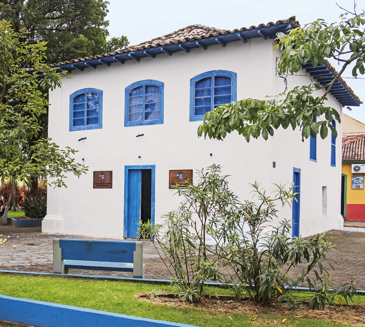 Fotografia. Fachada de uma casa de paredes brancas, janelas e porta azuis, e um telhado triangular, com algumas árvores ao redor.