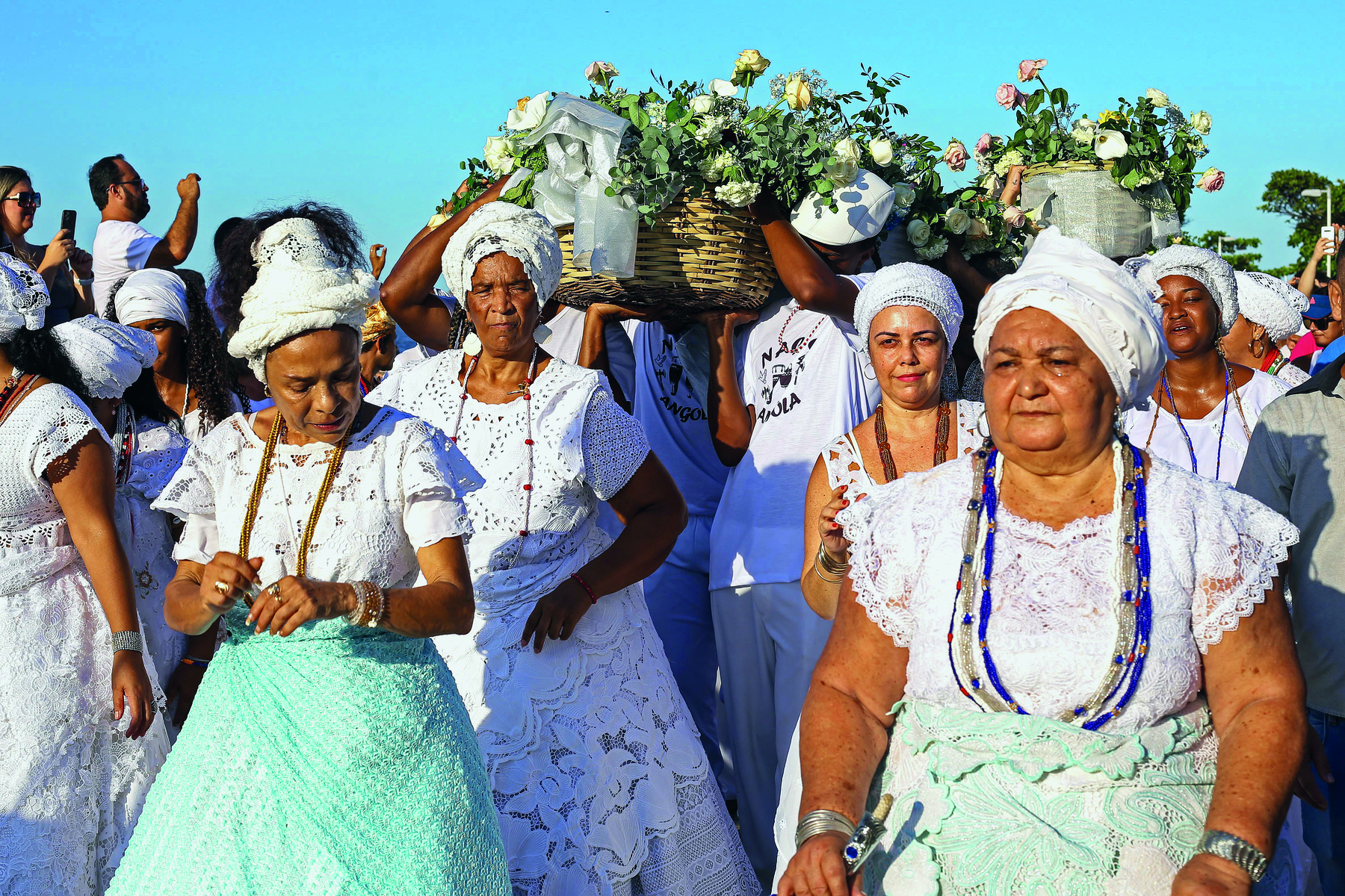 Fotografia. Em primeiro plano, um conjunto de mulheres lado a lado, trajando vestidos brancos, turbantes de mesma cor ao redor de suas cabeças e colares de cores diferentes. Ao centro, destaque para algumas pessoas carregando cestas com rosas brancas.
