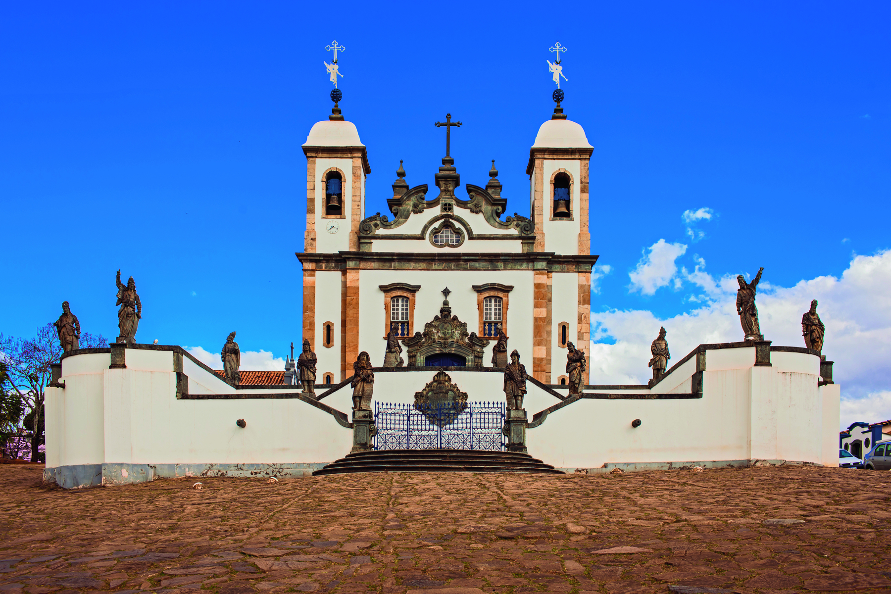 Fotografia. Fachada de uma igreja com paredes brancas, duas torres laterais com tetos em formato de cúpula, uma cruz central, muros a circundando e um portão frontal. Sobre os muros, diversas esculturas.