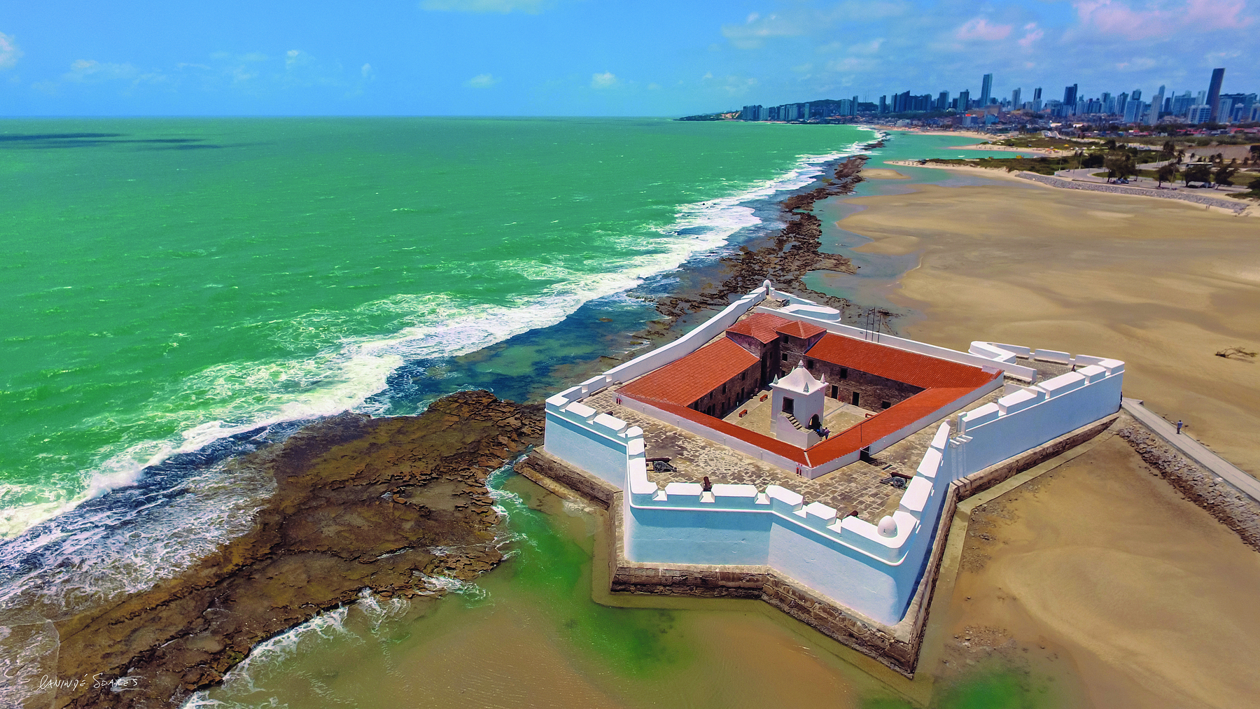 Fotografia. Vista aérea de uma fortaleza com paredes brancas, em formato de estrela com cinco pontas, com uma torre central, sobre um solo de areia, em uma praia banhada por águas esverdeadas.