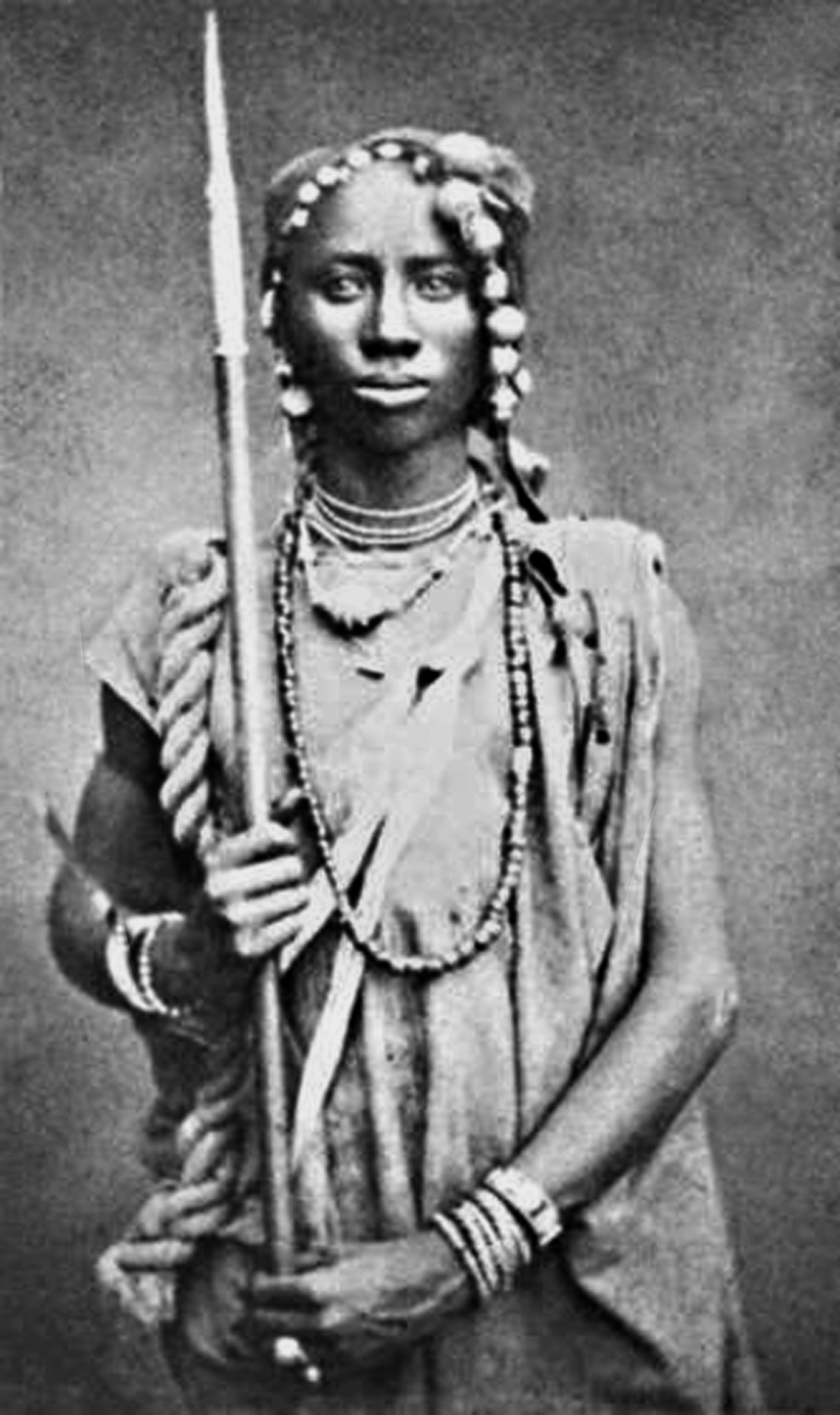 Fotografia em preto e branco. Uma mulher vista de frente, com cabelos escuros e trançados, vestindo uma camisa clara e colares sobre o peito e ao redor do pescoço, segurando uma lança com uma das mãos.