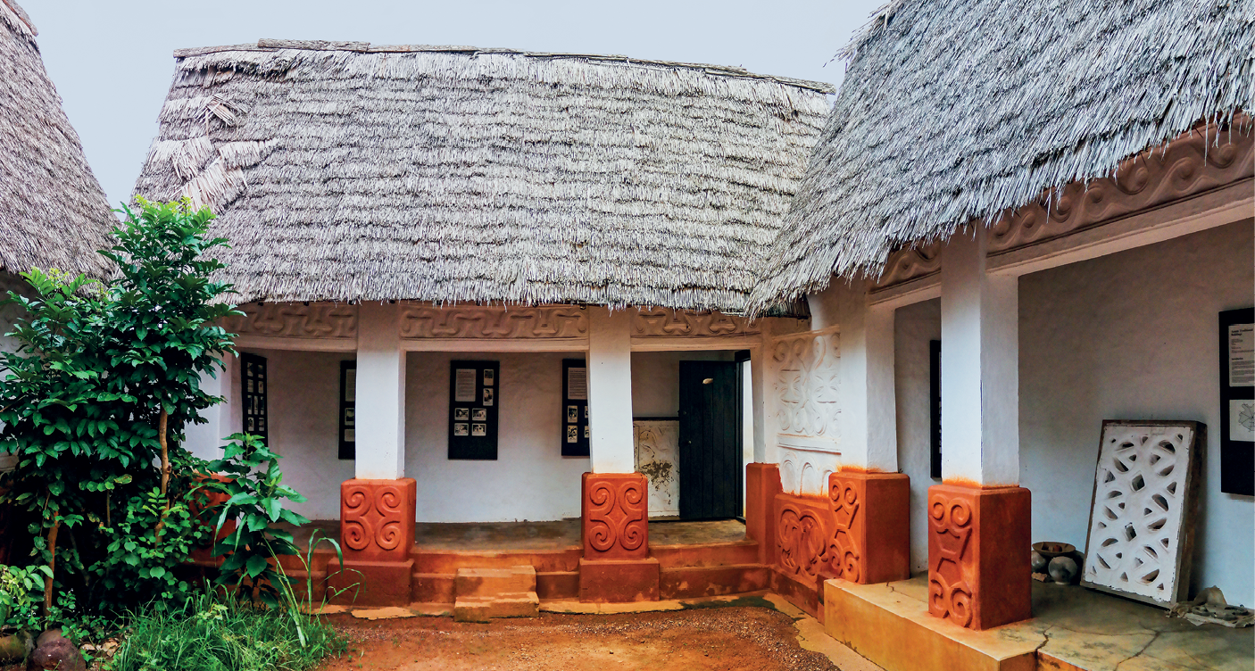 Fotografia. Vista da fachada de uma casa com paredes brancas, vigas verticais com bases laranja sustentando tetos triangulares cobertos por palha. As paredes possuem janelas e portas de madeira.