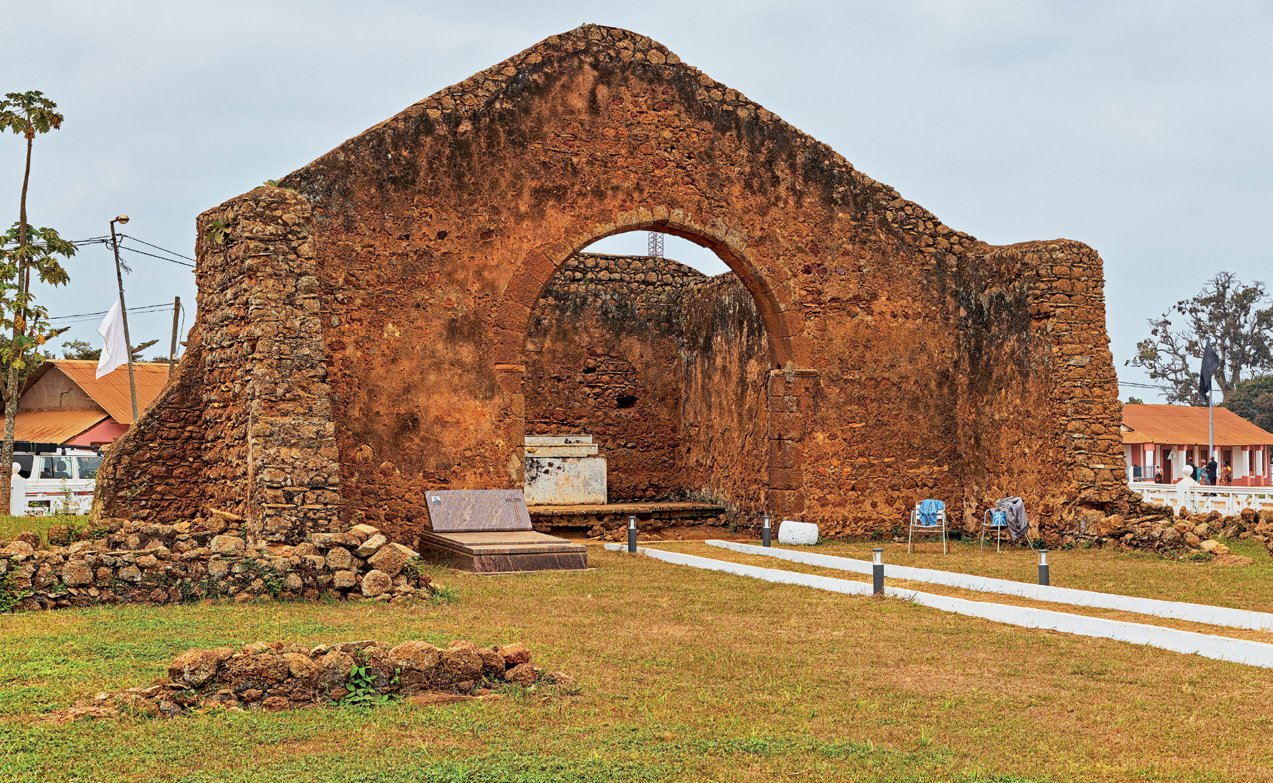 Fotografia. Vista da fachada de uma construção em ruínas, com paredes de pedra, formato triangular e uma abertura central em forma de arco, indicando um pequeno altar.