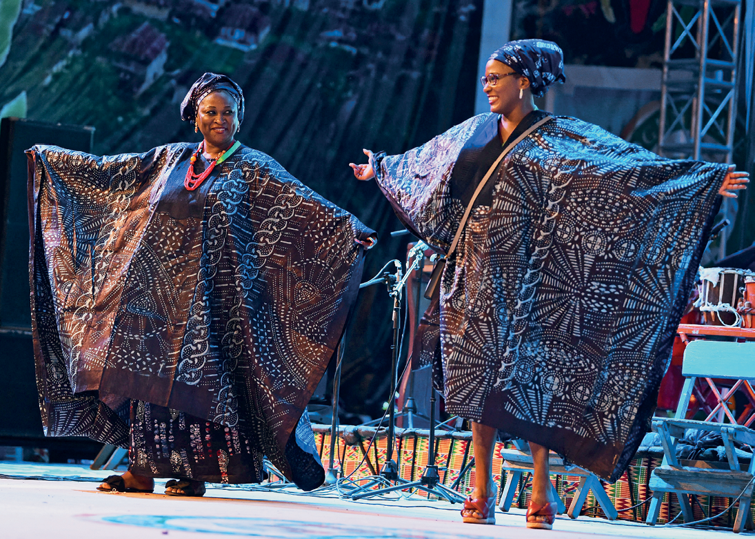 Fotografia. Duas mulheres, lado a lado, em pé sobre um palco, ambas trajando túnicas semelhantes, com cores escuras e detalhes formados por linhas e figuras geométricas.