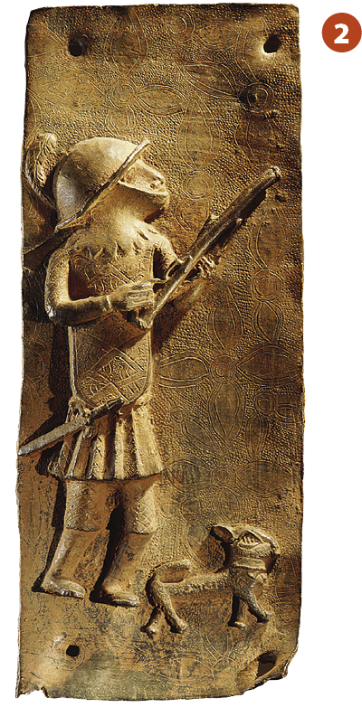Escultura sobre placa. Representação de um guerreiro visto de lado, em pé, vestindo uma armadura, botas e um capacete na cabeça. Ele segura um armamento com as duas mãos e possui uma faca atada à sua cintura. Aos seus pés há um cachorro.