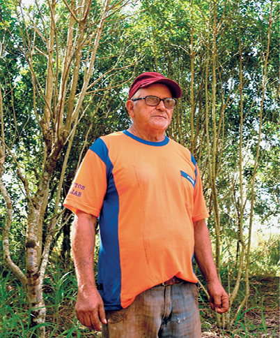 Fotografia. Um homem de cabelos e barba grisalhos, vestindo uma camiseta laranja, um boné vermelho sobre a cabeça e um par de óculos no rosto. Ao fundo, uma densa vegetação.