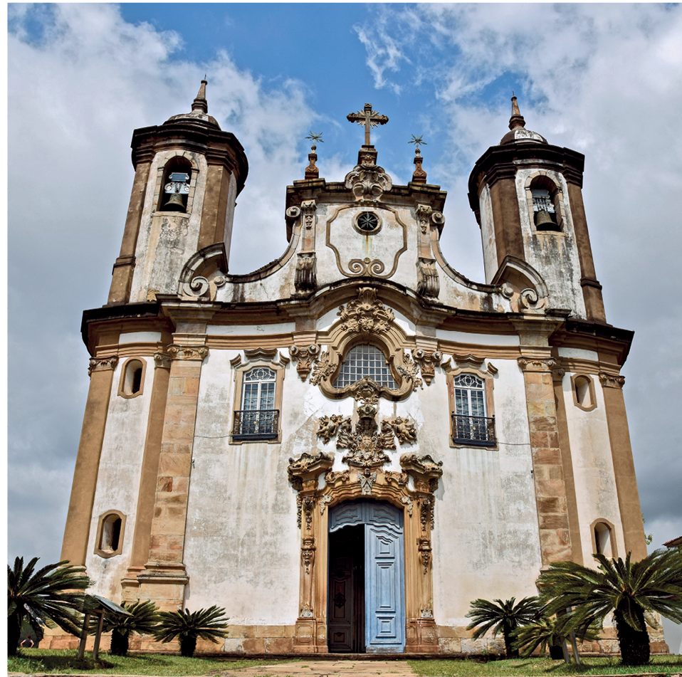 Fotografia. Fachada de uma igreja com paredes brancas e detalhes em tijolos, duas torres laterais, uma cruz, ao centro, e uma entrada frontal retangular.