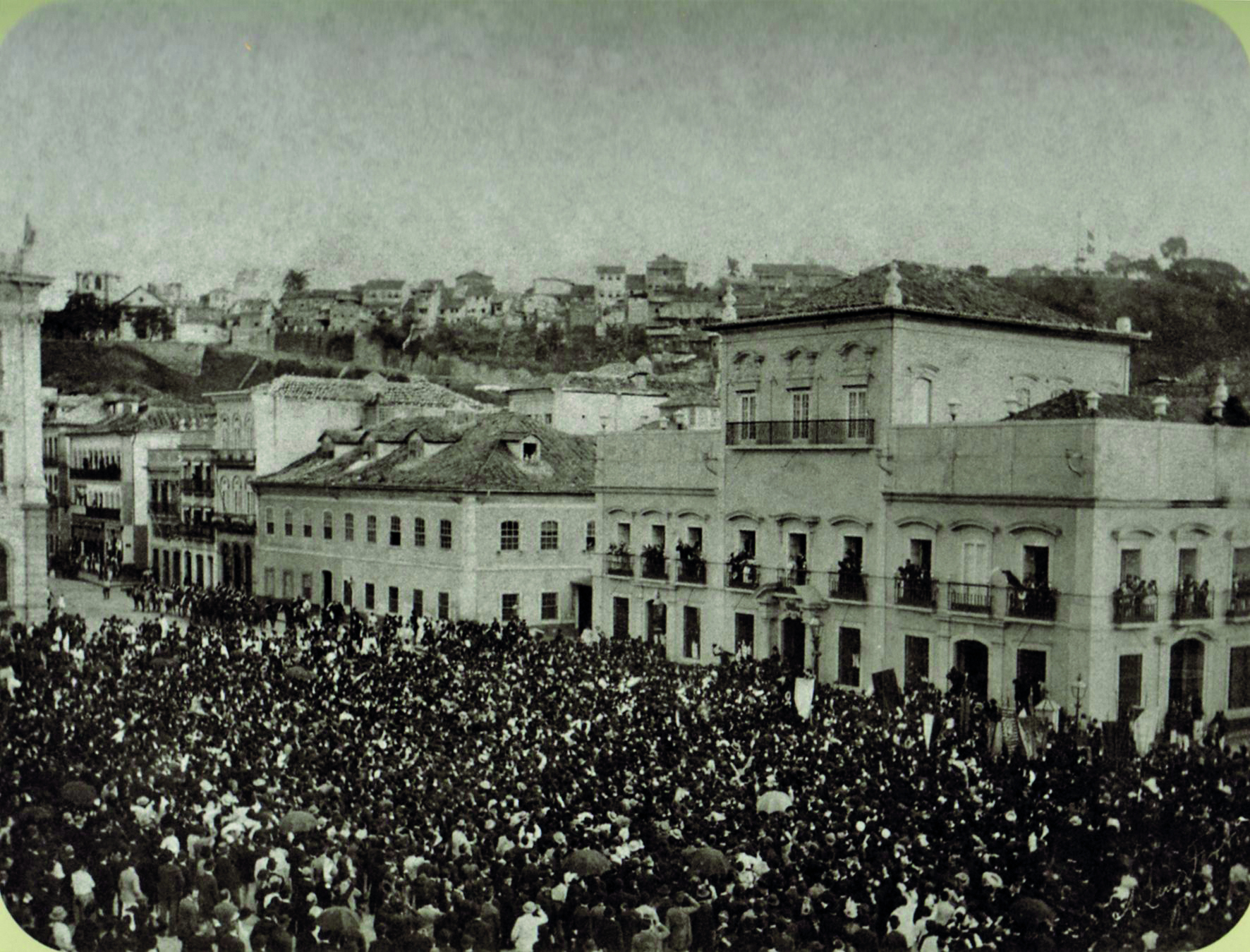 Fotografia em preto e branco. Uma multidão de pessoas aglomeradas em uma rua cercada por construções baixas de estilo colonial. Na porção direita da imagem, no Paço Imperial, pessoas ocupam os balcões das janelas e acenam para a multidão.