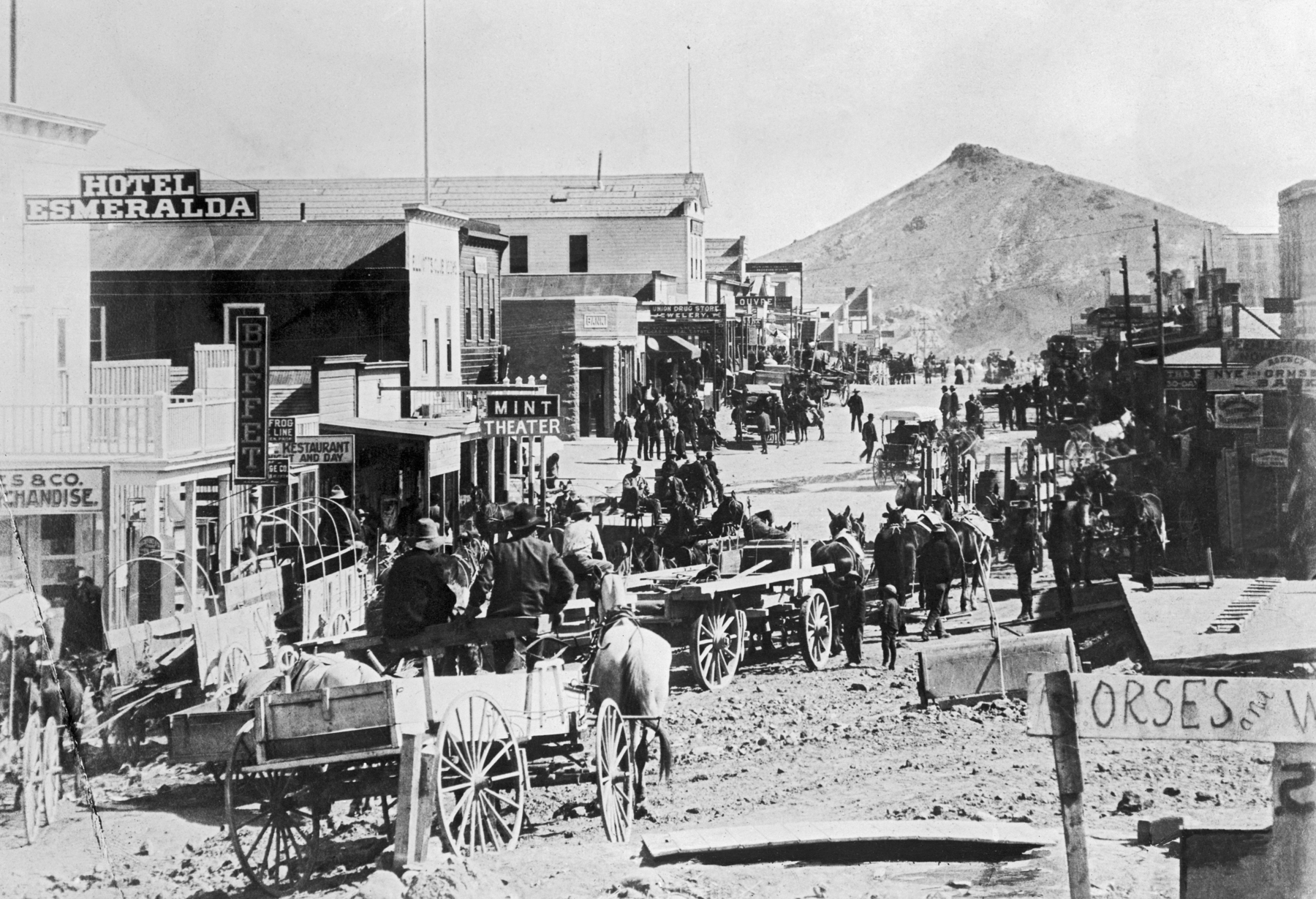 Fotografia em preto e branco. Em uma estrada de terra, cercada por algumas construções de madeira, homens em carroças puxadas por cavalos. Ao fundo, uma montanha alta.