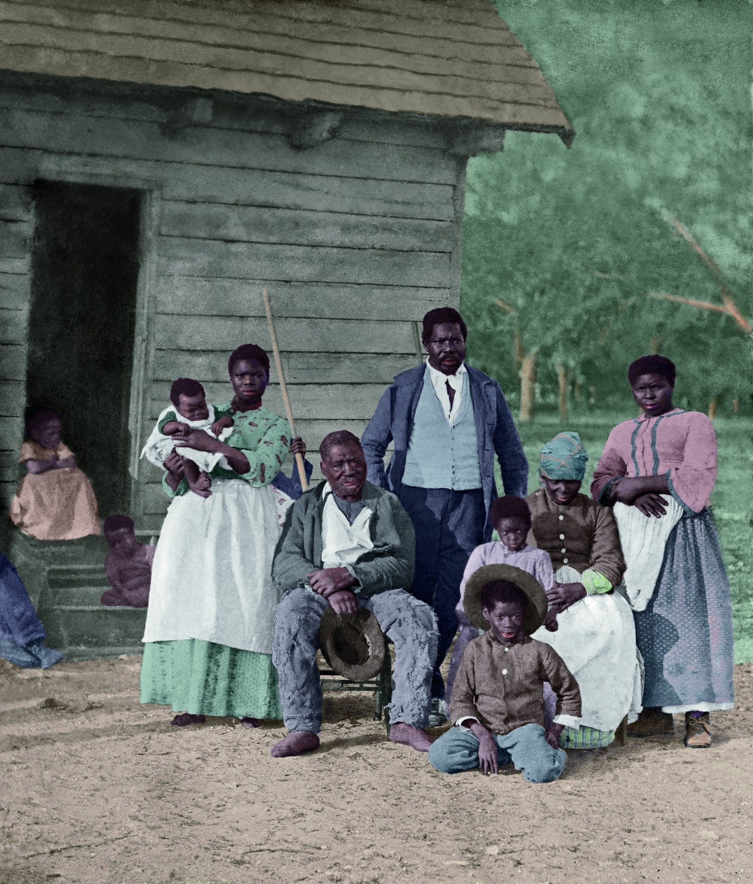 Fotografia. Um grupo de pessoas lado a lado, entre crianças, bebês, homens e mulheres, alguns em pé, alguns sentados, sobre um solo terroso. Ao fundo, uma casa de madeira.