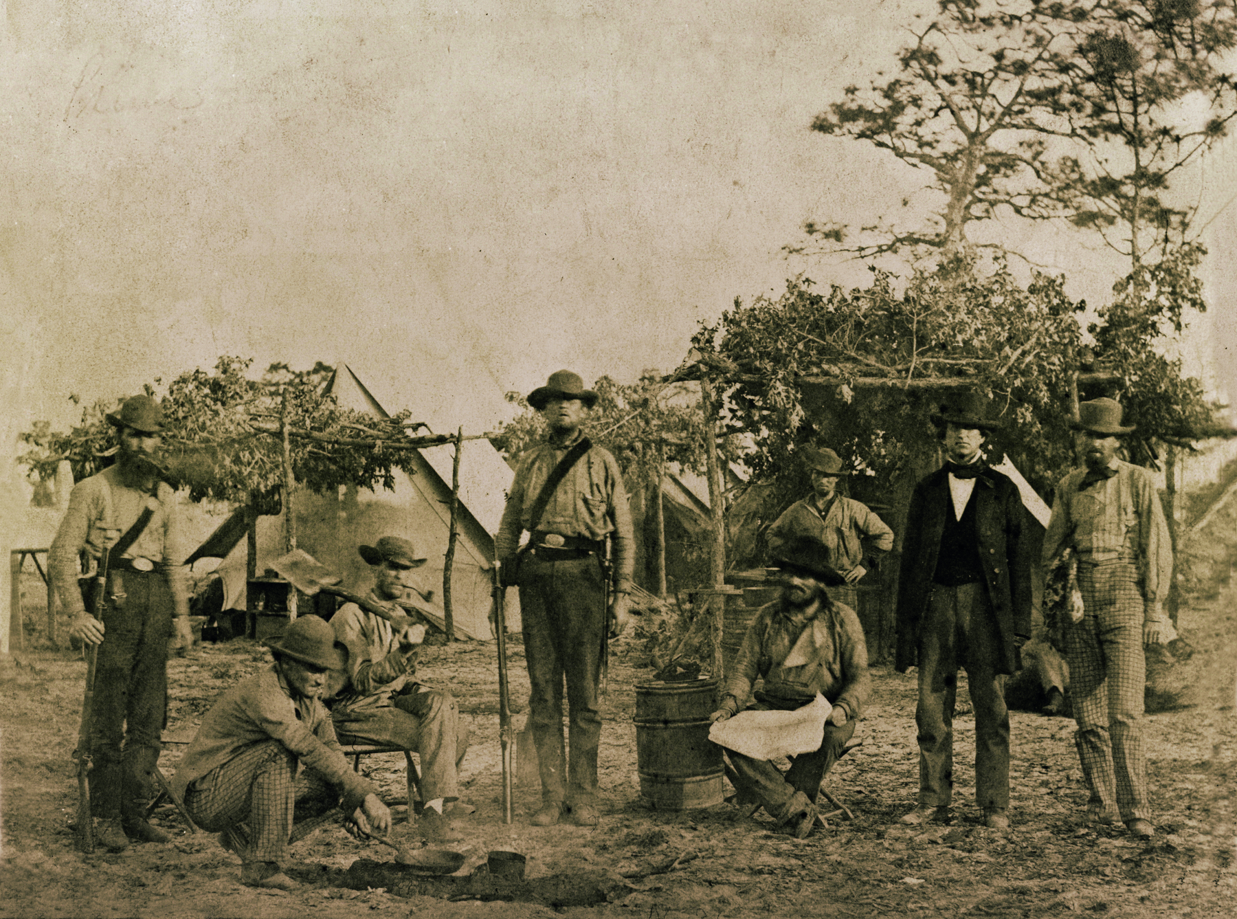 Fotografia em preto e branco. Homens lado a lado, uns em pé, outros agachados no solo, alguns portando armas de fogo. Em segundo plano, árvores e algumas pequenas cabanas.