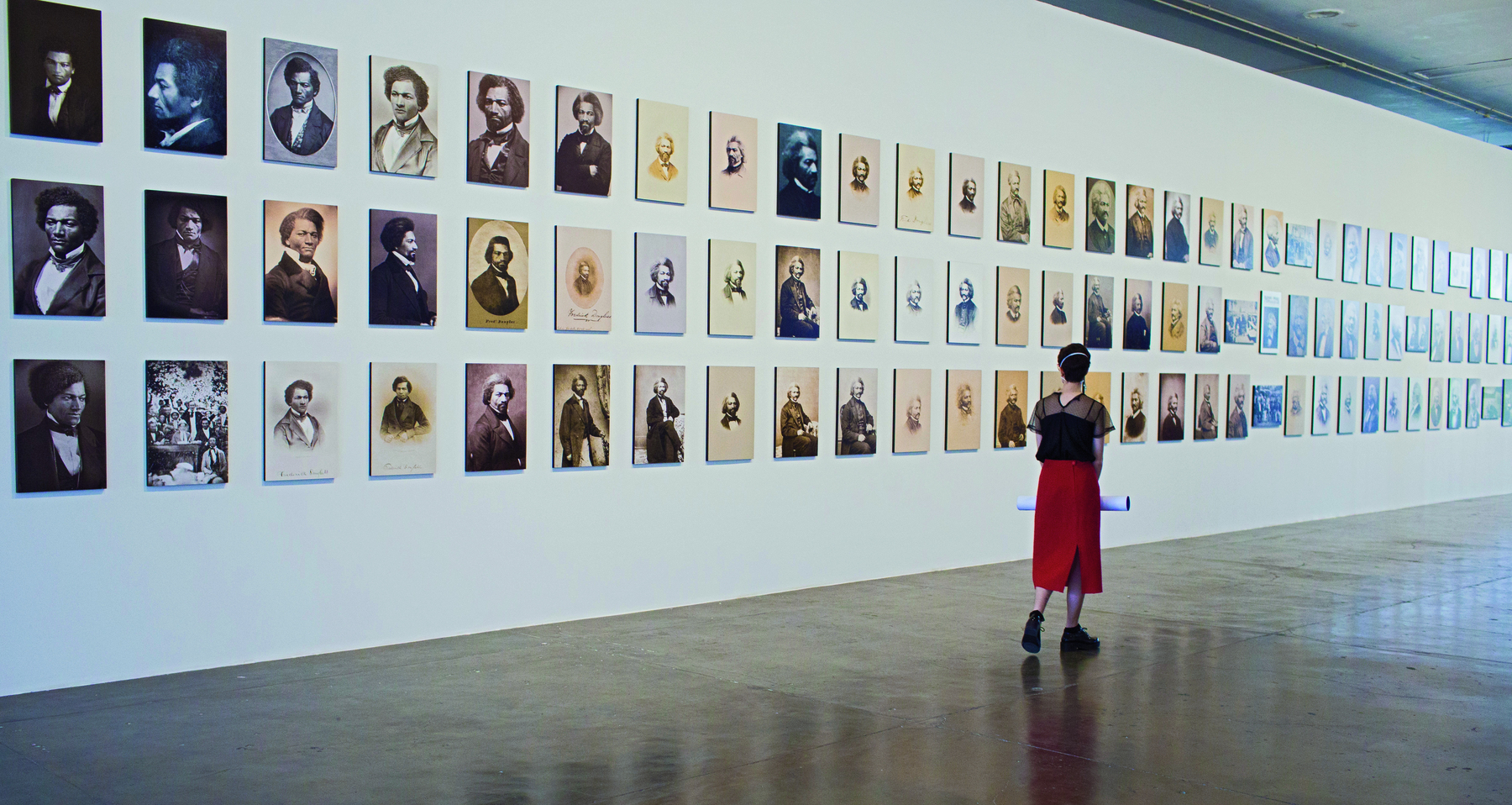 Fotografia. Vista de costas, uma mulher em pé, vestindo uma blusa preta e uma longa saia vermelha, a frente de uma parede branca contendo diversos quadros com diferentes retratos de um mesmo homem em exposição.