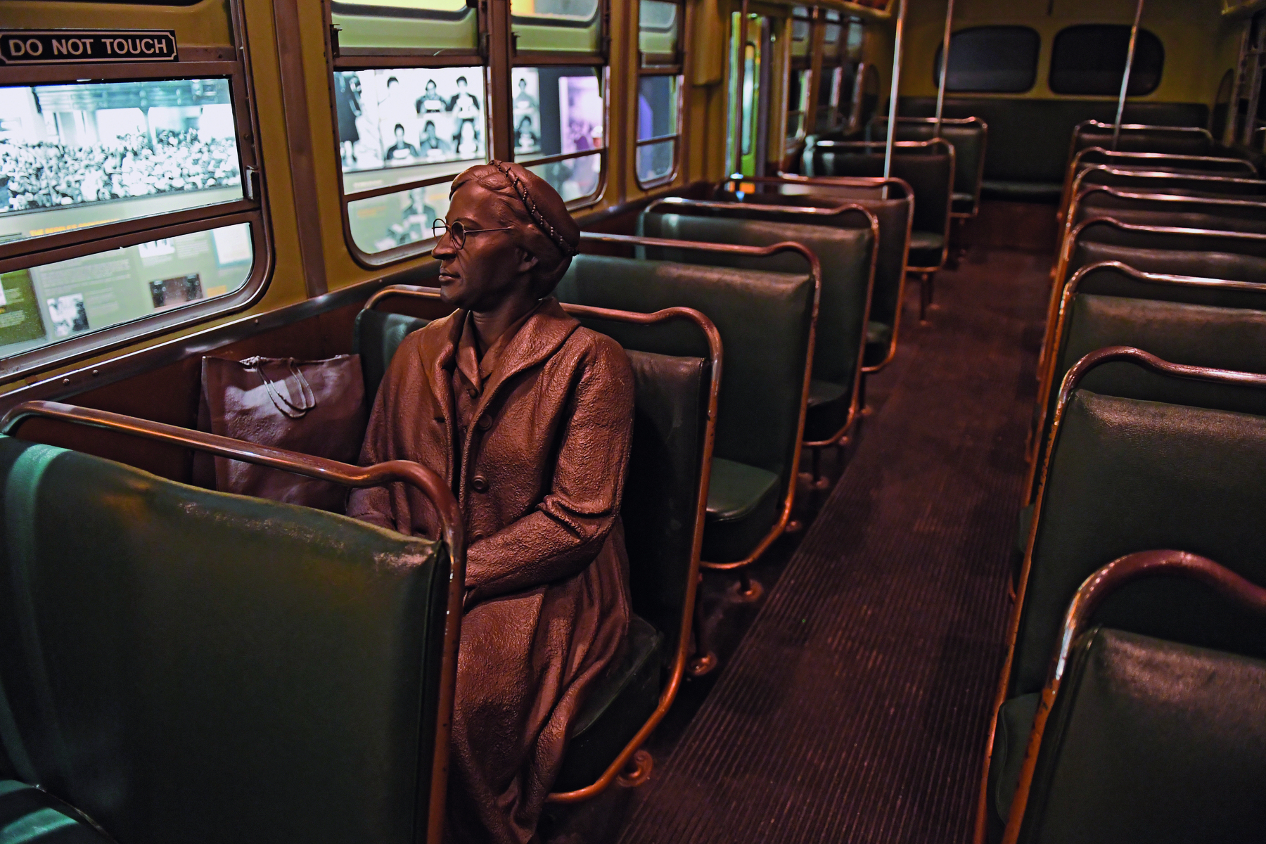 Fotografia. Escultura feita de metal representando uma mulher sentada sobre um banco de um ônibus. A escultura está dentro de um ônibus antigo, com bancos para duas pessoas com estofado verde escuro e piso de madeira escura.