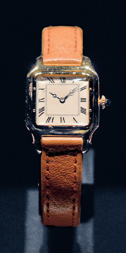 Fotografia.  Sobre um fundo azul, um relógio de pulso em formato quadrado, com ponteiros prateados, números romanos e pulseira de couro marrom.