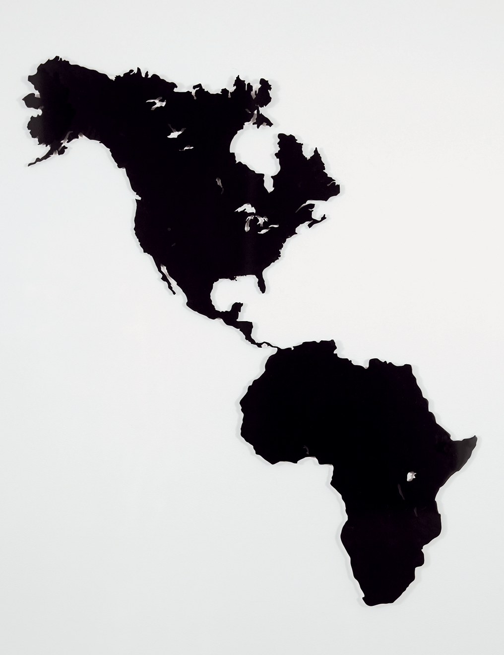 Fotografia. Obra em preto e branco. Sobre um fundo branco, em preto, um mapa com os territórios da América do Norte e Central, conectados com o continente africano (em lugar do que habitualmente seria a América do Sul).