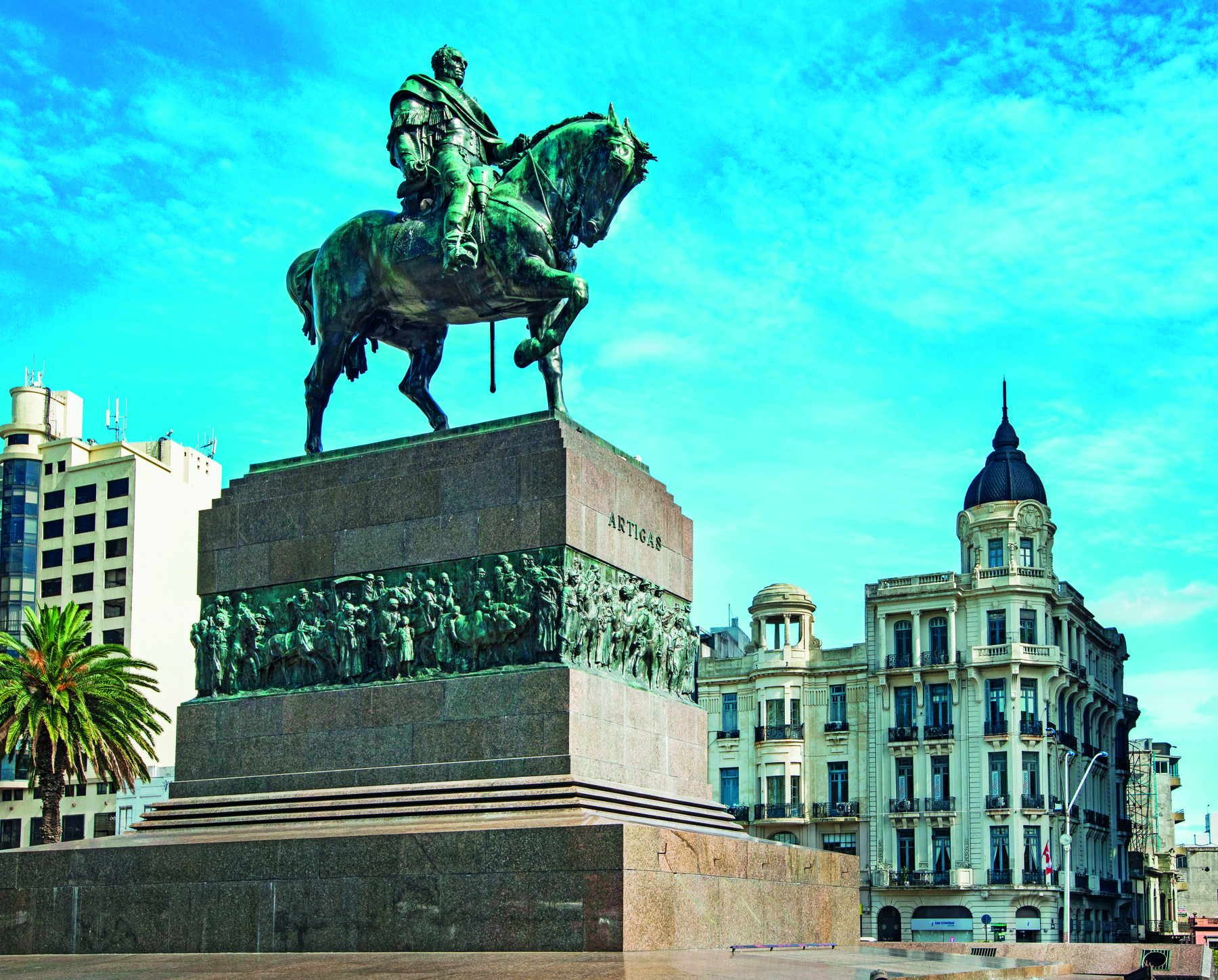 Fotografia. Sobre um pedestal de pedra, a estátua de um homem vestindo uma armadura, montado em um cavalo. Em segundo plano, edifícios, um deles com uma cúpula azul. No pedestal está gravado o nome: Artigas.