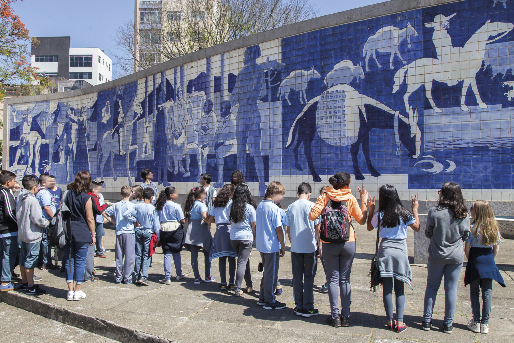 Fotografia. Um grupo de crianças lado a lado, em frente a um mural de azulejos, representando homens e animais em atividades diversas.