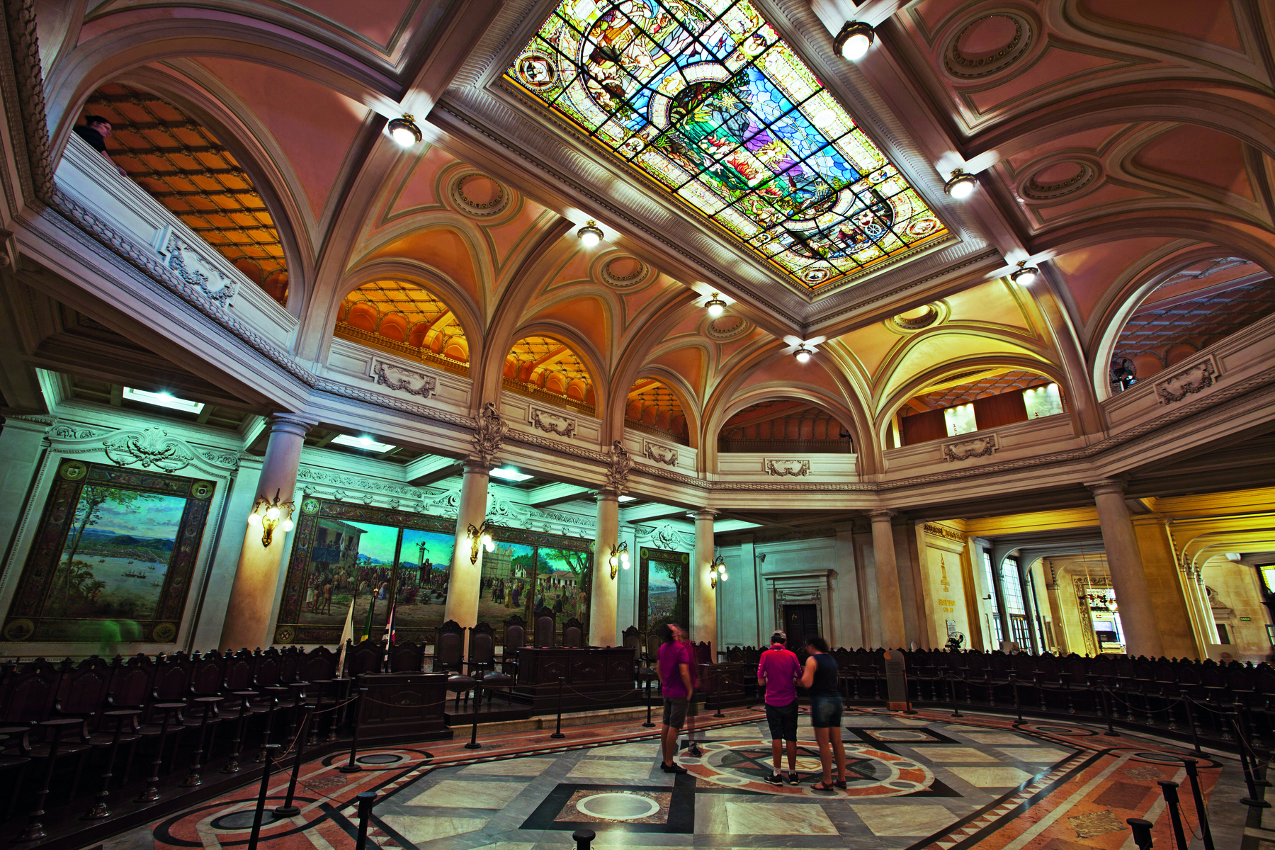 Fotografia. Um amplo salão, com teto em cúpula, contendo vitrais coloridos, e pelas paredes, entre colunas que terminam em arcos, pinturas e quadros expostos. No meio do salão, pessoas em pé agrupadas.