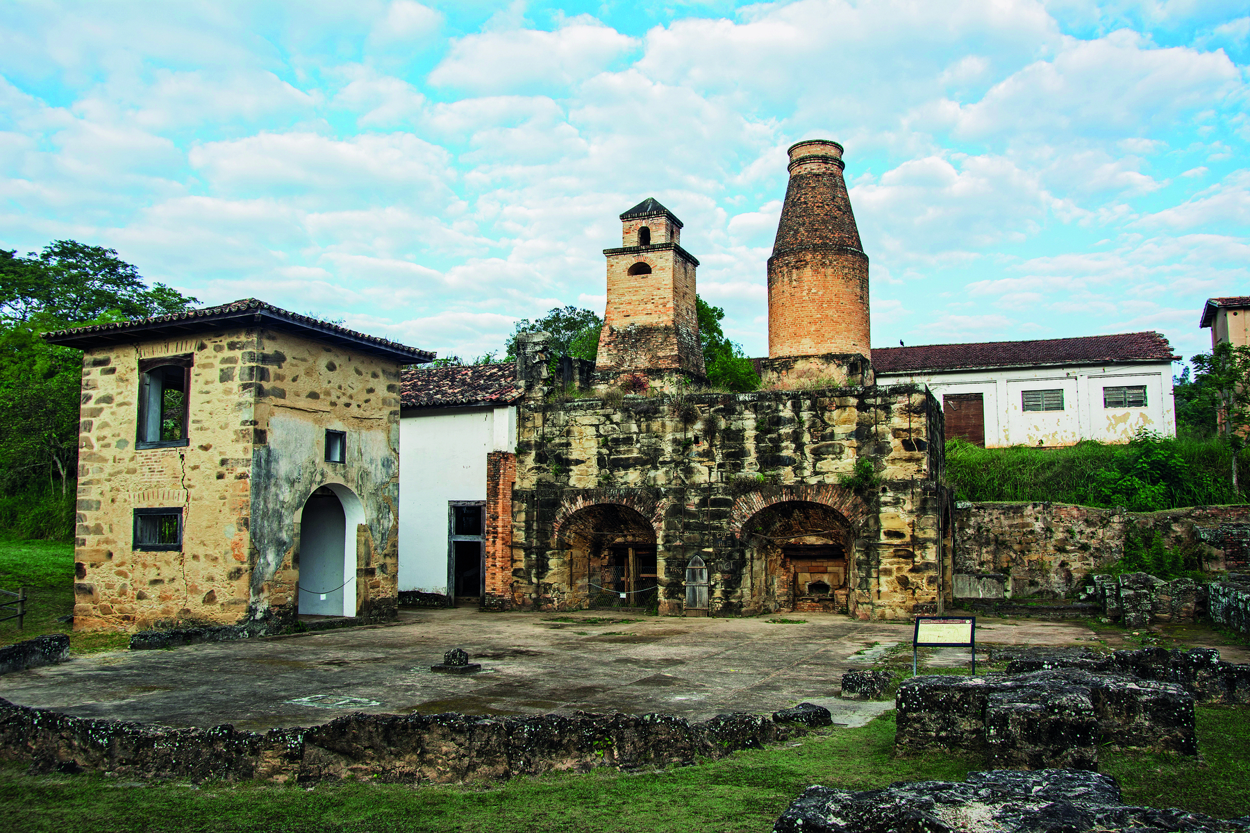 Fotografia. Vista frontal das ruínas uma pequena fábrica com paredes de tijolos, dois fornos, duas chaminés ao centro, e um pequeno pátio plano à frente.