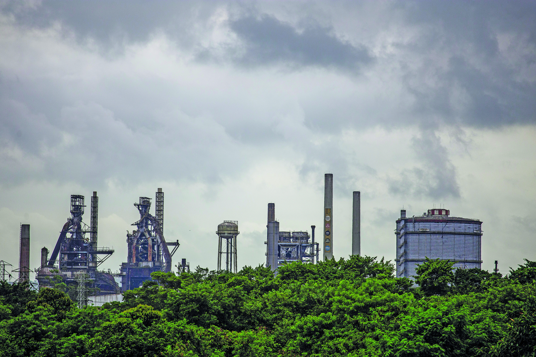 Fotografia. Sob um céu escuro e nublado, vista de chaminés de fábricas e usinas, lado a lado. Em primeiro plano uma faixa de vegetação formada por copas de árvores.
