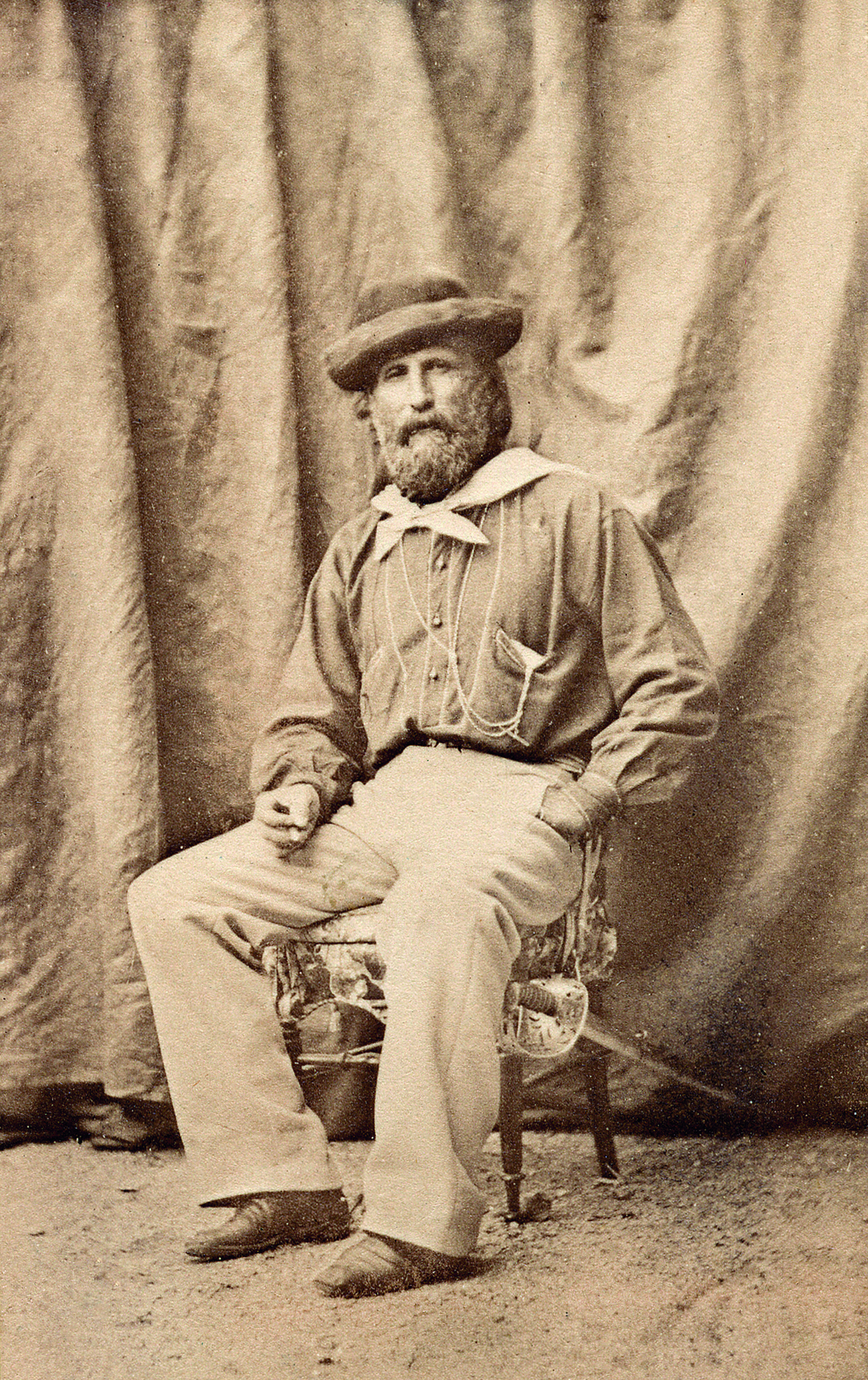 Fotografia em preto e branco. Um homem com barba volumosa, vestindo uma camisa, lenço claro amarrado no pescoço com um nó, calça e um chapéu com abas, sentado sobre uma cadeira. A mão esquerda está no bolso e a direita está sobre a sua perna.