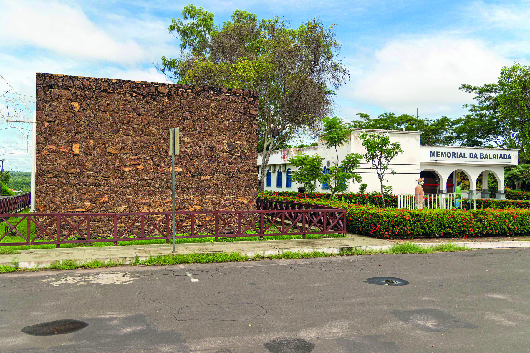 Fotografia. Uma parede de pedra em ruínas, cercada por uma pequena grade, e à direita, uma casa com paredes brancas, entrada composta por três arcos, contendo o texto sobre sua fachada: 'Memorial da Balaiada'.