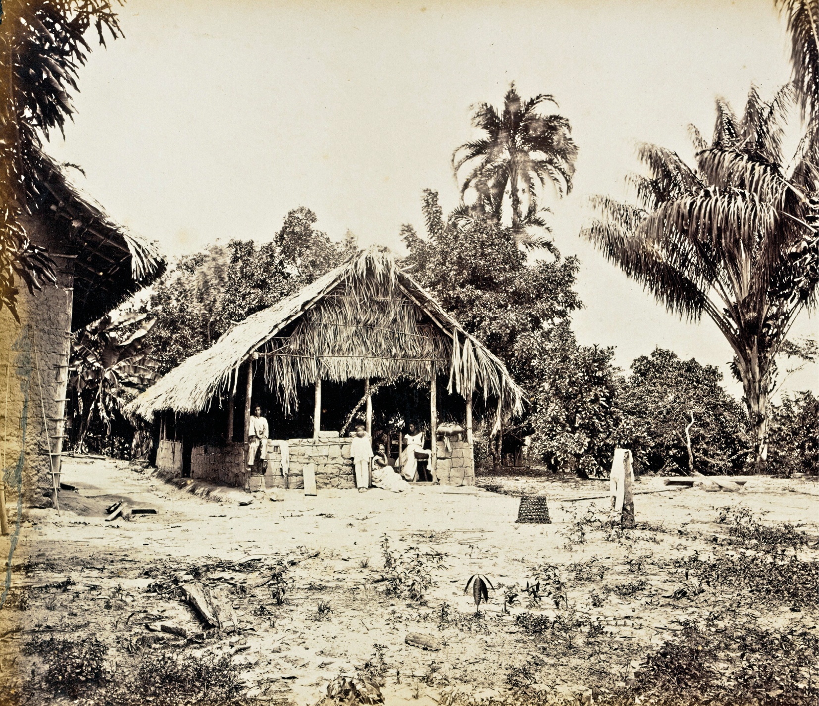 Fotografia em preto e branco. Uma cabana feita de folhagens, estacas e barro, em um terreno plano, com alguns arbustos. Ao fundo, árvores e palmeiras.
