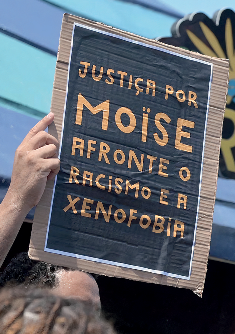 Fotografia. Detalhe de uma imagem com destaque para a mão de um homem segurando um cartaz feito de papelão, com o texto: 'Justiça por Moïse afronte o racismo e a xenofobia'.
