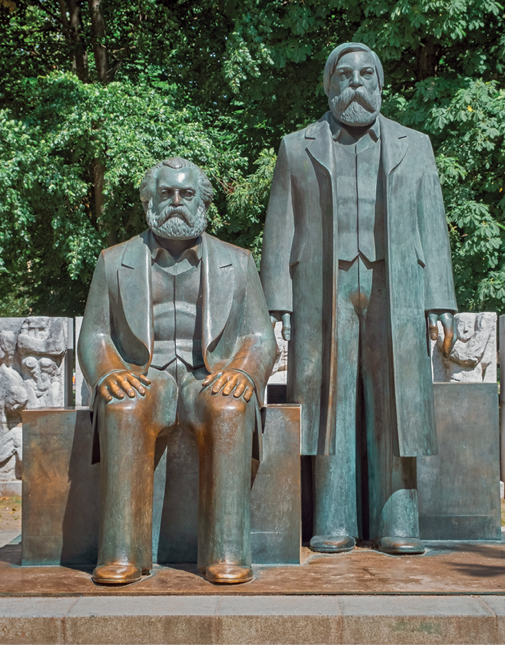 Fotografia. Monumento com a estátua de dois homens, lado a lado, ambos vestindo casacos, coletes e calças compridas, com cabelos curtos, bigodes e barbas volumosas. Um deles, à esquerda, está sentado. Outro, à direita, está em pé.