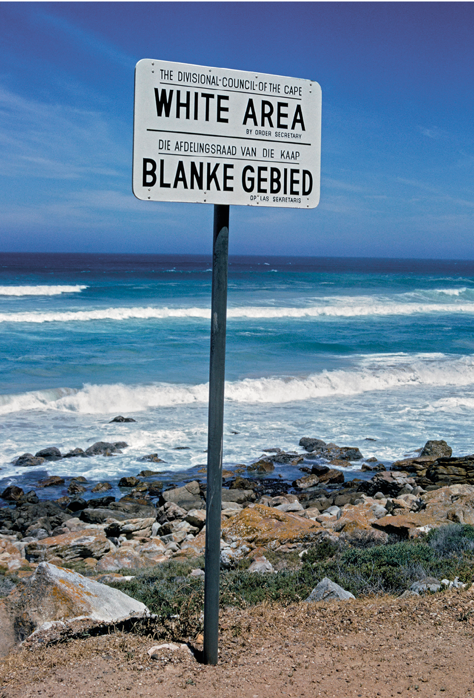 Fotografia. Em uma praia, fincada sobre uma porção de areia, com pedras às margens de águas marítimas azuis, em destaque, uma placa na cor branca com o texto: 'Área branca', na parte superior, em inglês, na parte inferior, em africâner.