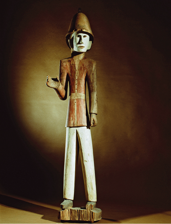 Escultura. Sob um fundo escuro iluminado, uma escultura de madeira representando um homem em pé, vestindo um casaco vermelho com botões na frente, uma calça branca e um capacete escuro sobre sua cabeça.  Um dos braços dele está abaixado, ao longo do corpo; o outro, flexionado. Seu rosto está pintado na cor branca.