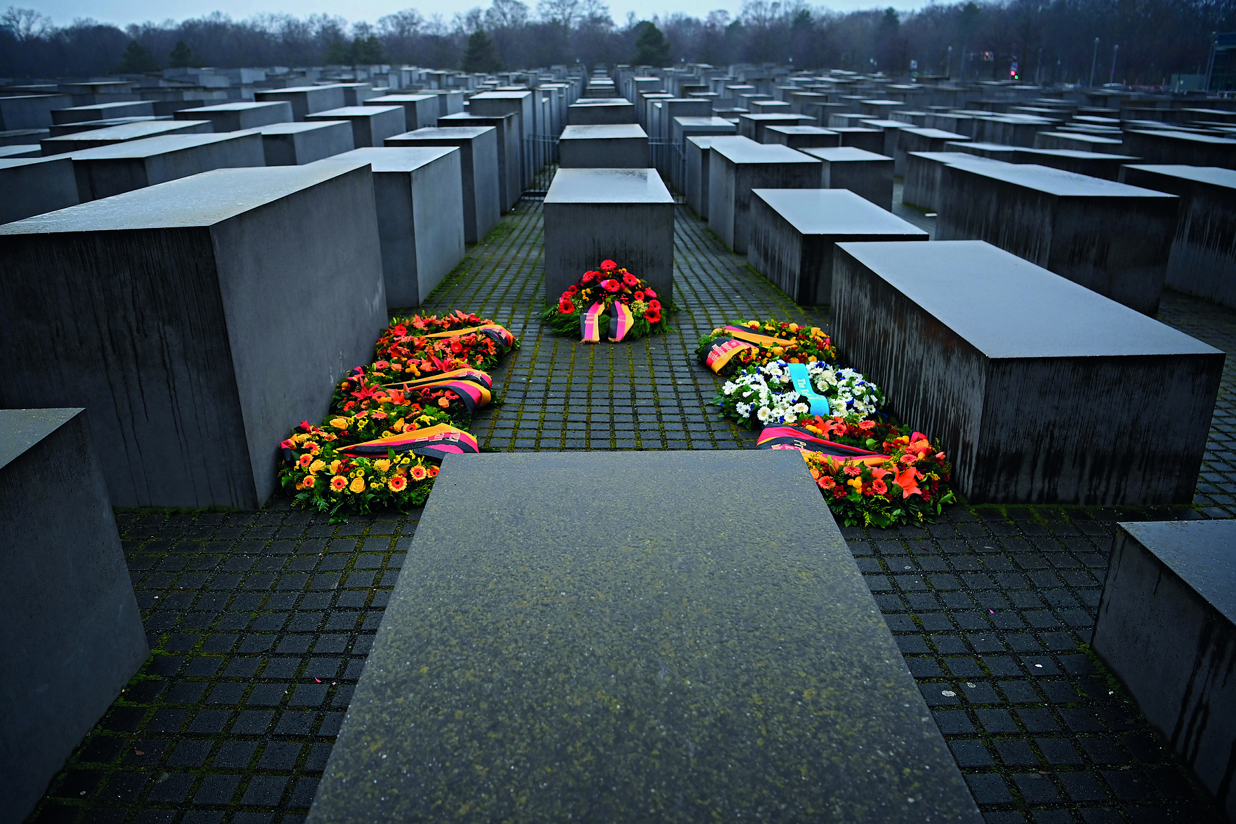 Fotografia. Vista de um memorial com diversos monumentos escuros, retangulares, de diferentes alturas, e ao centro, várias coroas de flores coloridas.