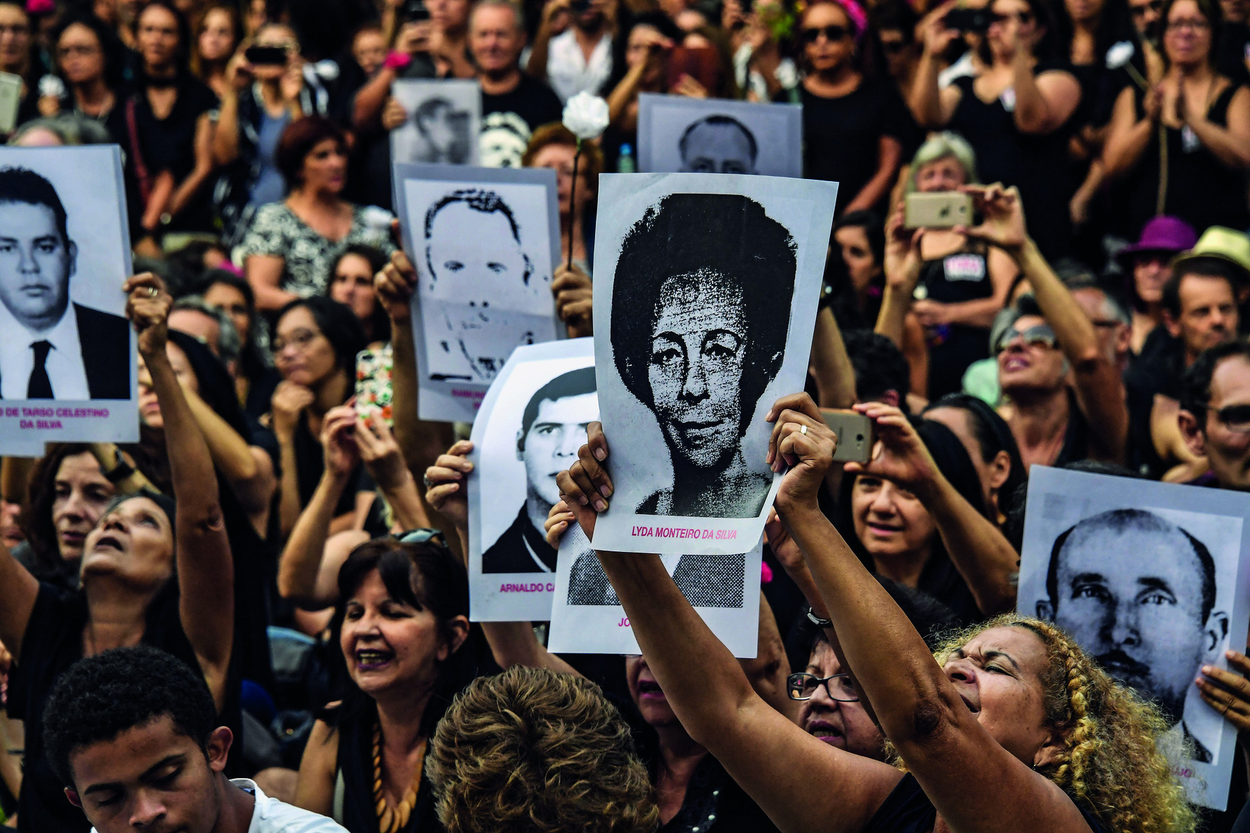 Fotografia. Uma multidão em uma manifestação. Com os braços erguidos para o alto, algumas pessoas seguram cartazes com retratos em preto e branco de diferentes pessoas, homens e mulheres. Os nomes dessas pessoas, ilegíveis na fotografia, estão escritos logo abaixo dos retratos, em caracteres na cor vermelha.