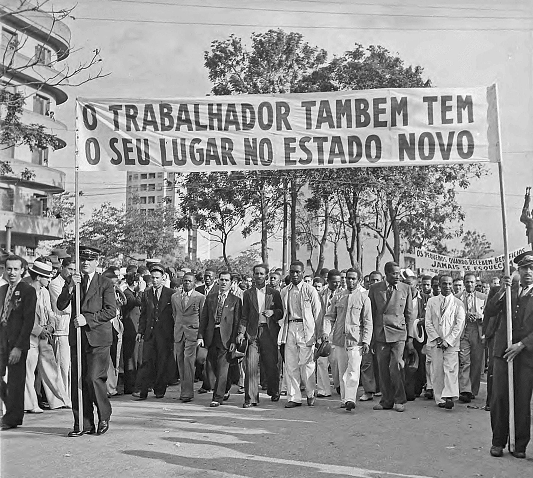 Fotografia em preto e branco. Um grupo de homens aglomerados, lado a lado, vestindo trajes sociais e portando uma faixa com o texto: 'O trabalhador também tem o seu lugar no Estado Novo'.