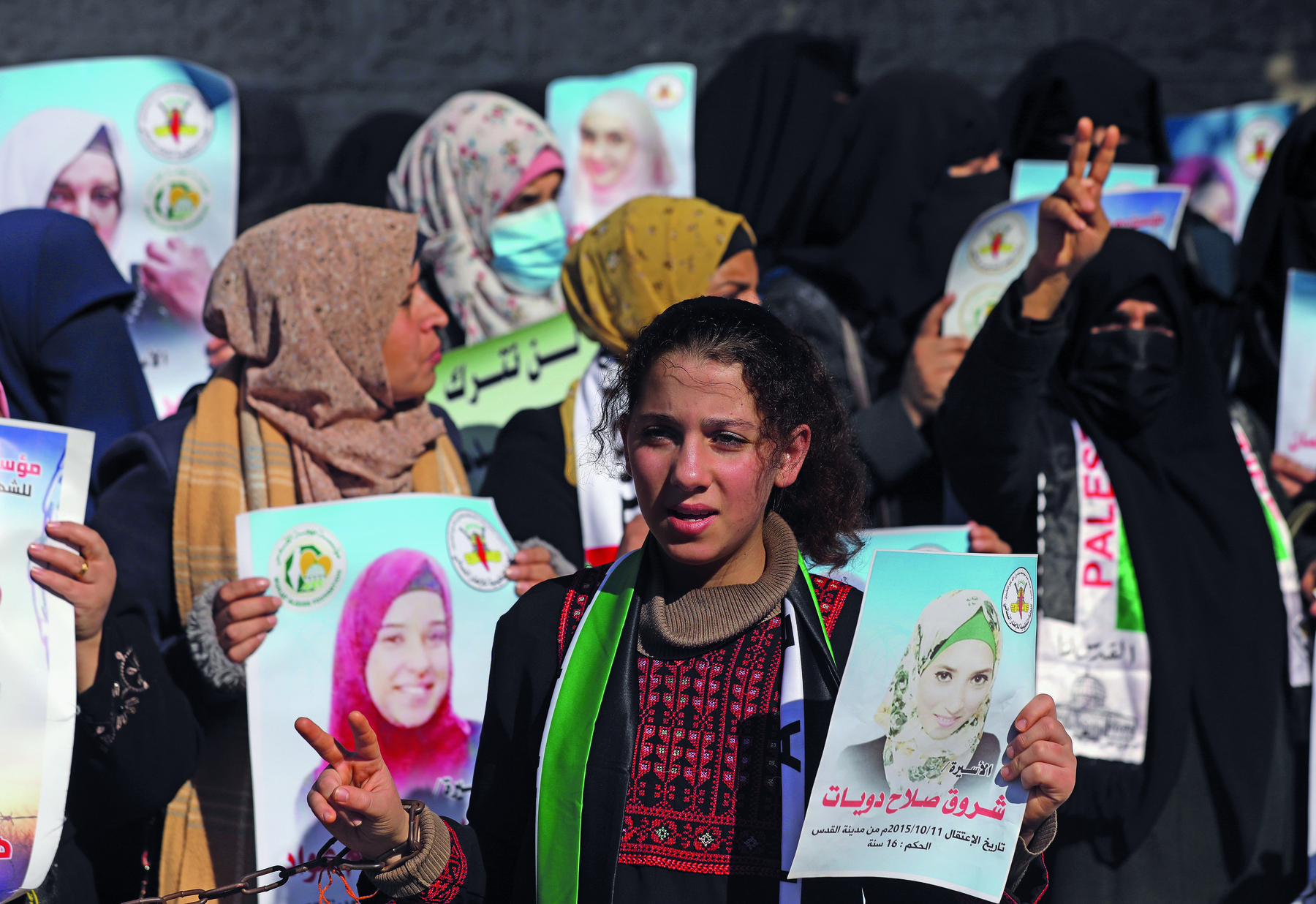 Fotografia. Um grupo de mulheres reunidas portando cartazes estampados com os rostos de outras mulheres. A maioria delas está usando vestes tradicionais que cobrem pescoço, orelhas e cabelo. Algumas cobrem também parte do rosto.
