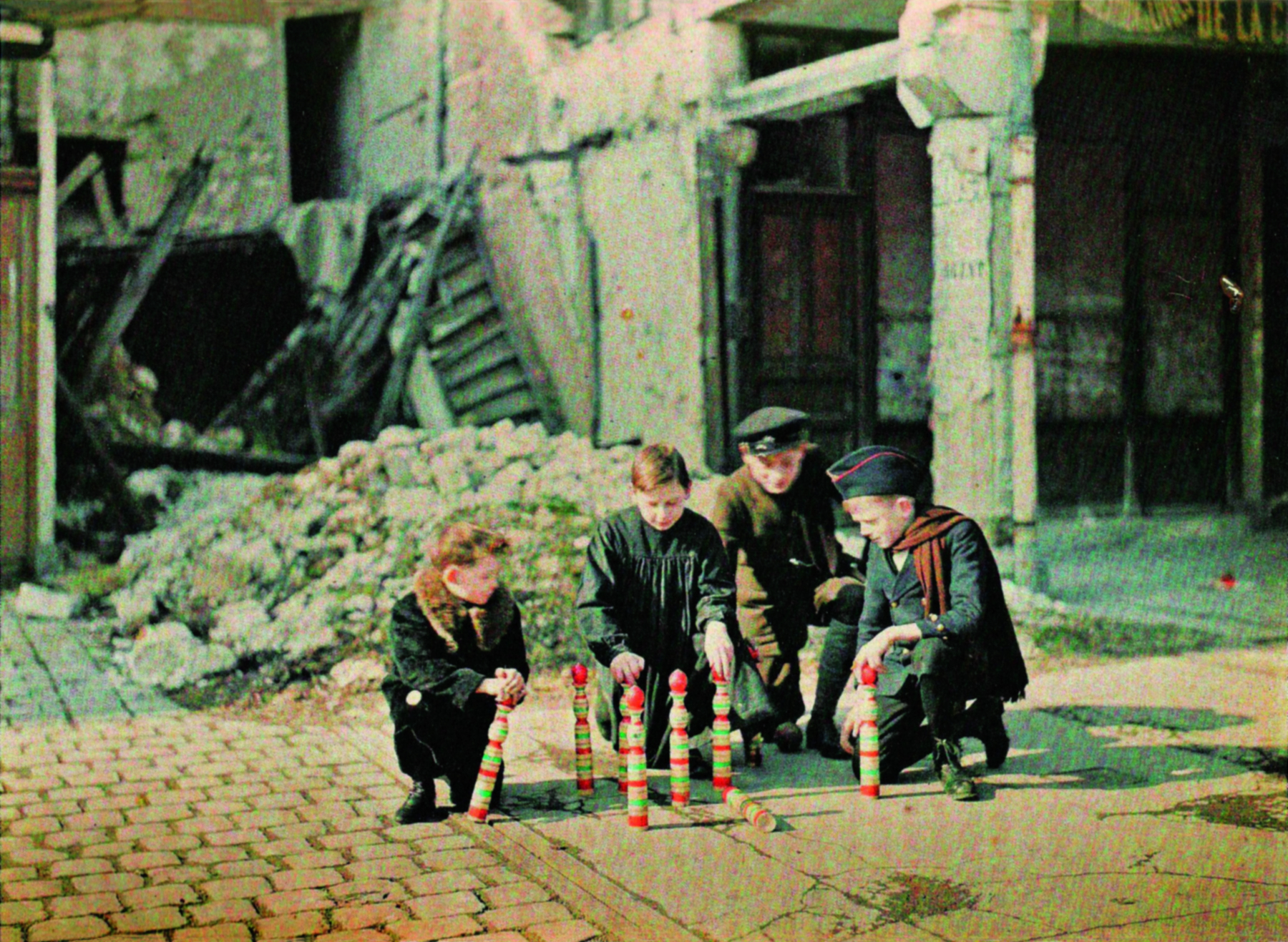 Fotografia. No centro, um grupo de meninos agachados ao redor de um brinquedo com torres verticais coloridas. Em segundo plano, os destroços de uma construção.
