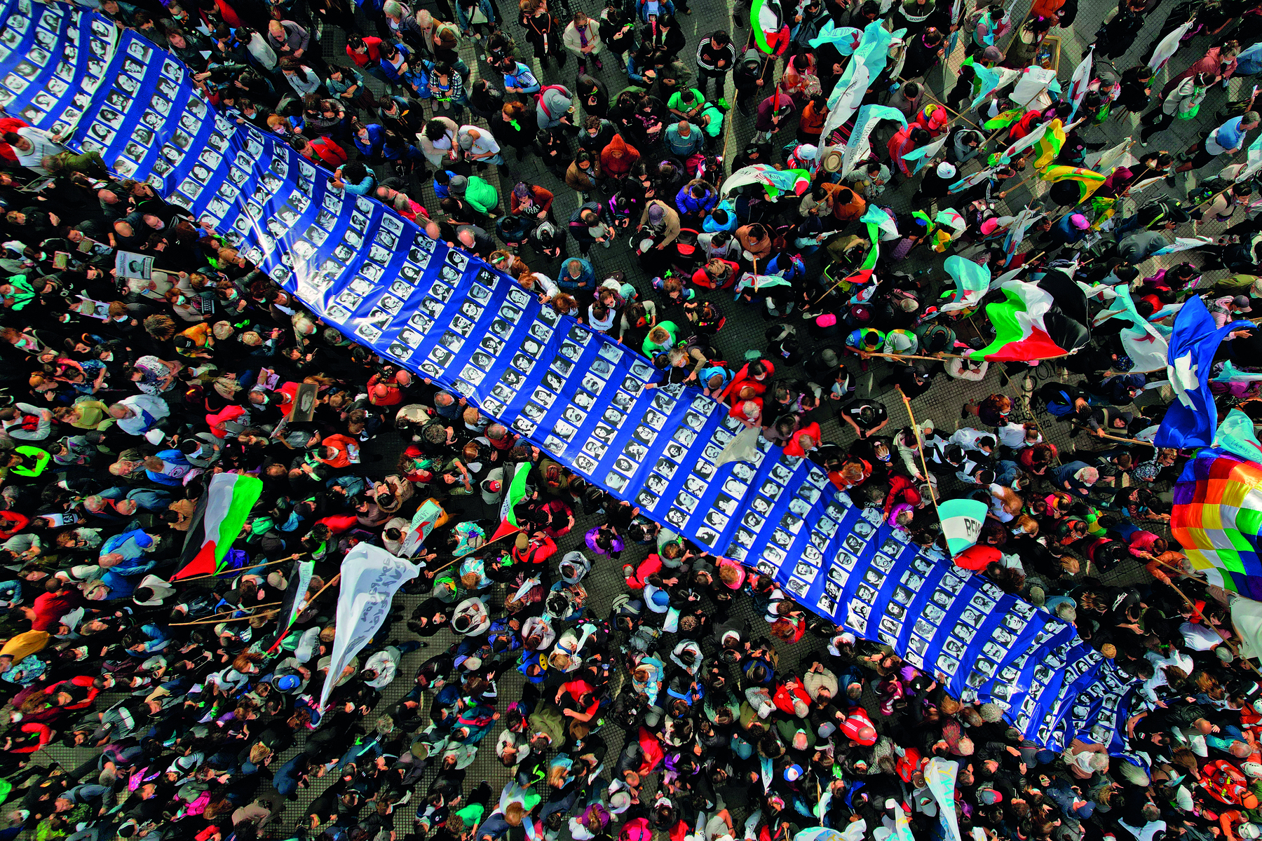 Fotografia. Vista aérea de uma multidão de pessoas em manifestação, portando faixas e bandeiras. No centro da imagem, uma longa faixa na cor azul com fotografias em preto e branco com retratos de centenas de pessoas.