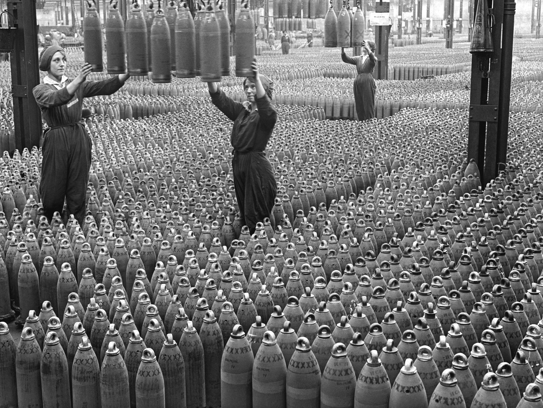 Fotografia em preto e branco. Em uma fábrica, três mulheres vestindo macacões escuros estão em pé, inspecionando mísseis que estão suspensos. O chão ao redor delas está coberto por mísseis enfileirados.