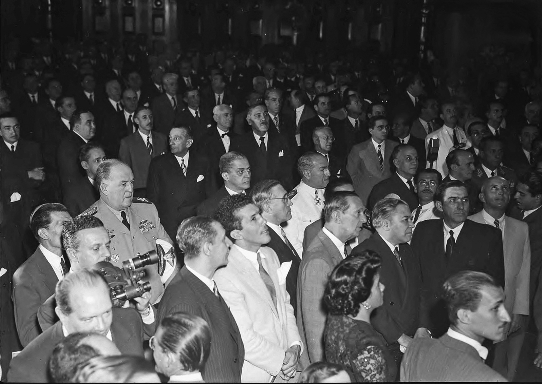 Fotografia em preto e branco. Um grupo de homens aglomerados em um salão, a maioria vestindo paletós escuros. No canto inferior direito, nota-se a presença de uma única mulher, de perfil.