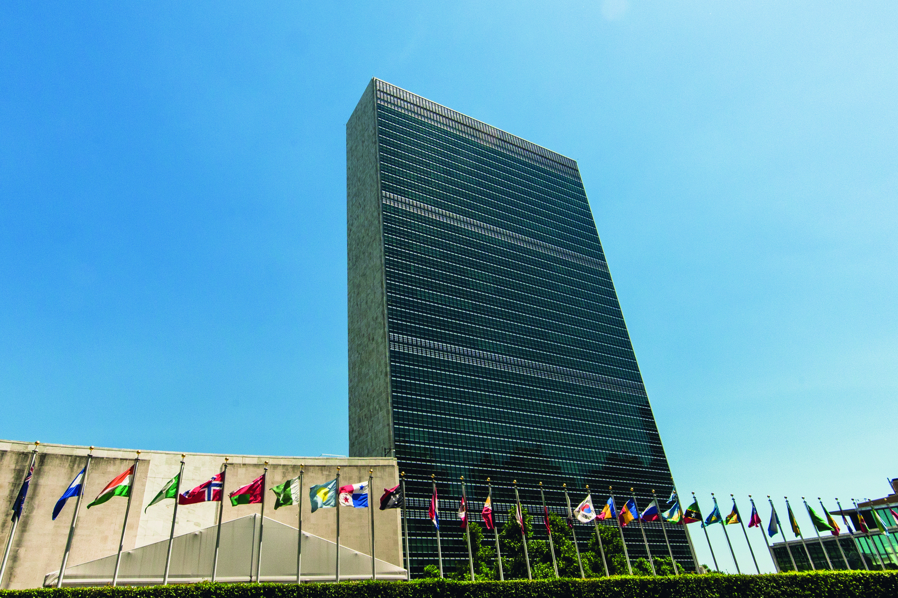 Fotografia. Fachada de um edifício alto, à sua frente, bandeiras hasteadas de diversos países.