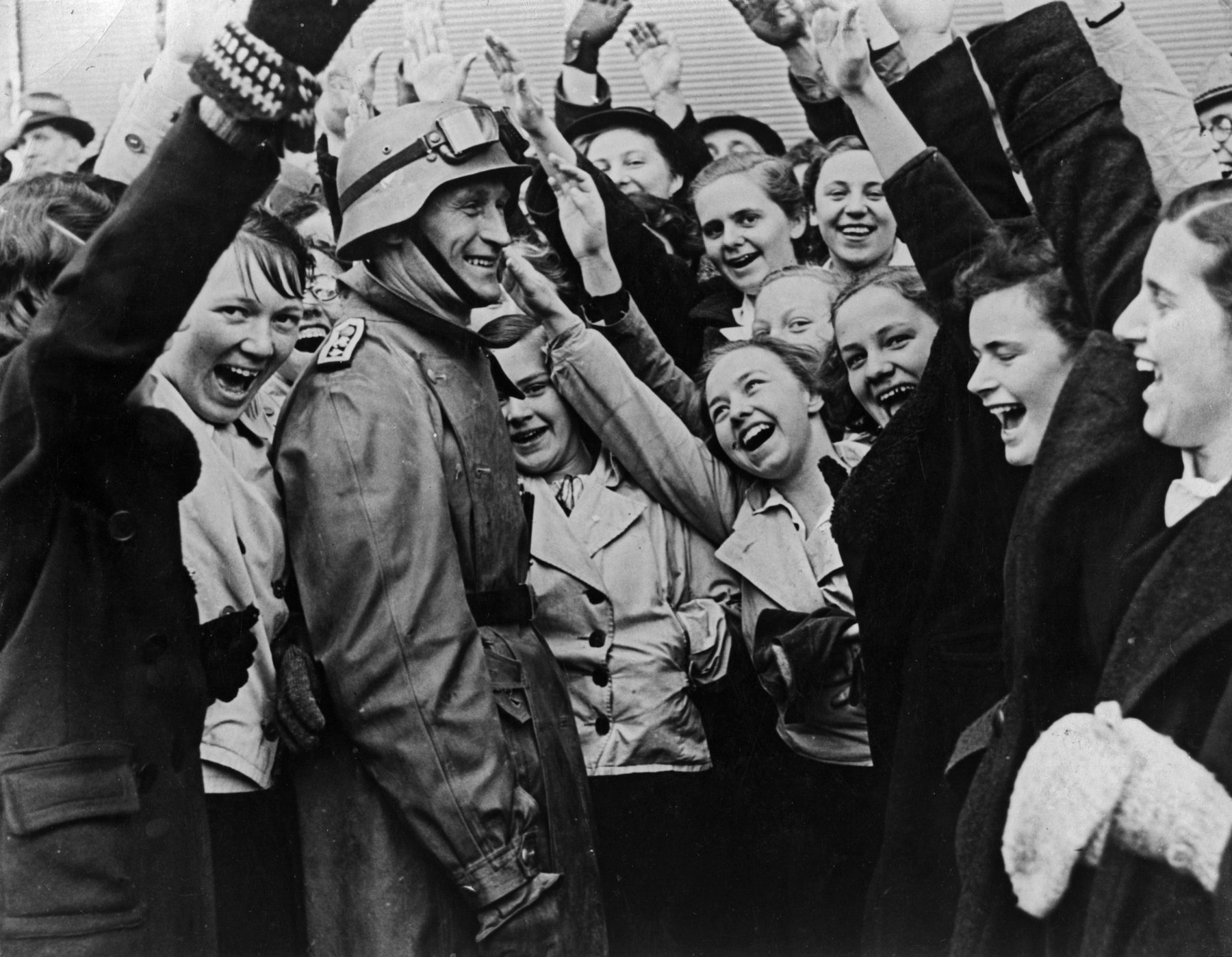 Fotografia em preto e branco. Um grupo de mulheres aglomeradas, com uma das mãos para frente, e os dedos esticados. No centro, um soldado vestindo uma farda e um capacete na cabeça.