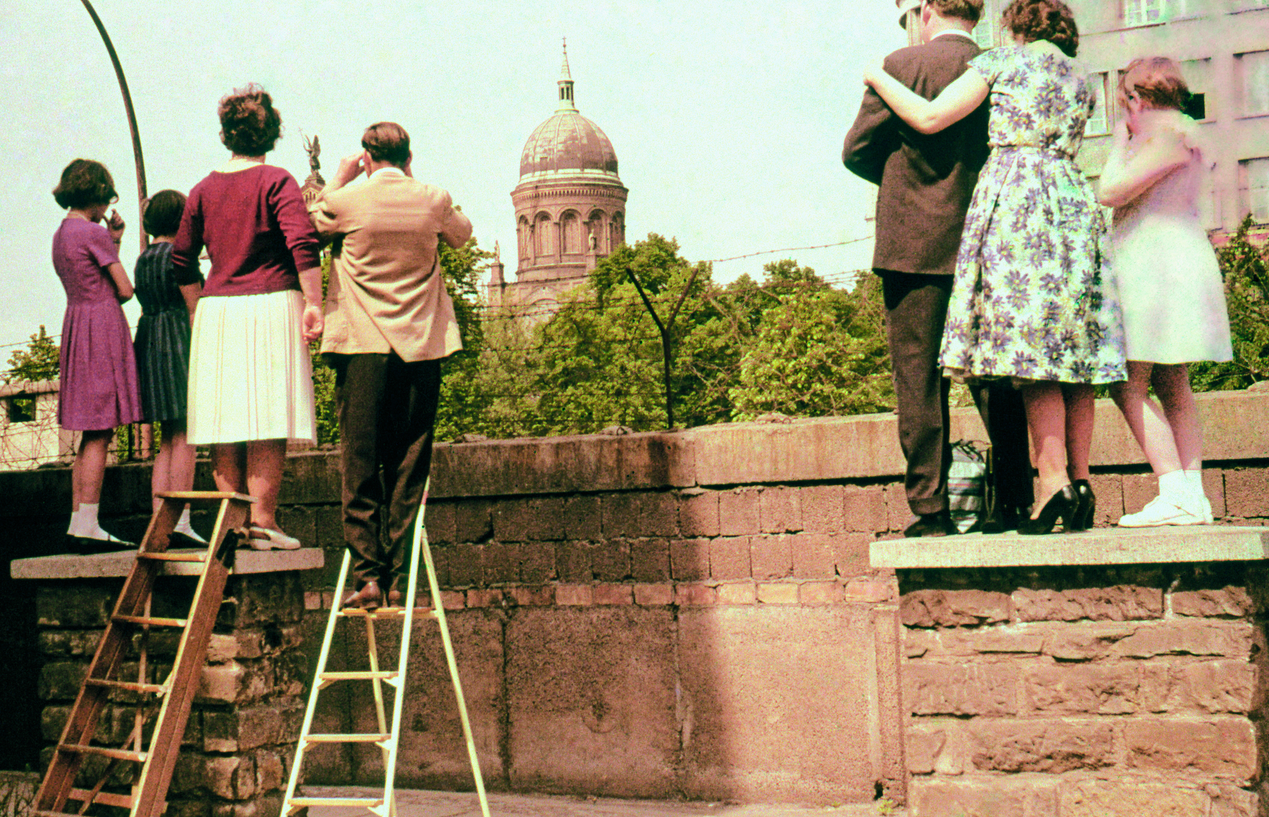 Fotografia. Um grupo de pessoas vistas de costas, em pé sobre muretas e escadas, observa o que há para além de um muro de tijolos. Em segundo plano, uma construção com uma cúpula de formato arredondado.