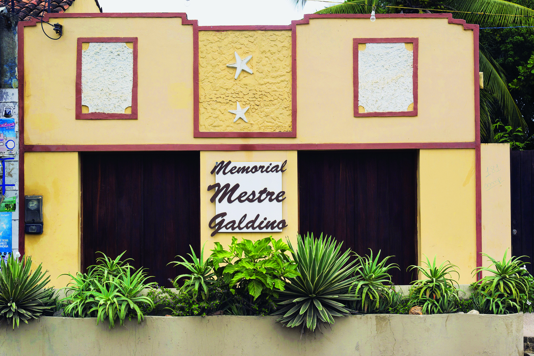 Fotografia. Vista da fachada central de uma pequena casa, com paredes amarelas, duas entradas laterais marrons, diversas plantas no solo, enfileiradas, e o texto: 'Memorial Mestre Galdino'.