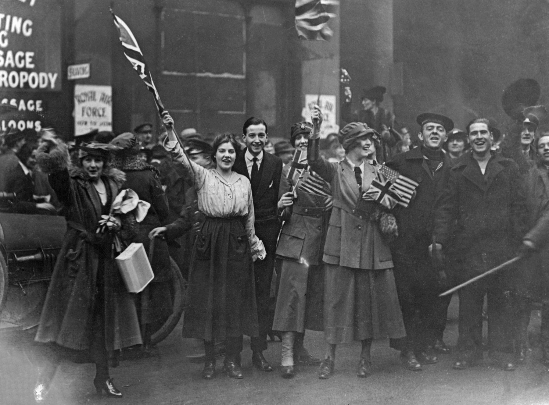 Fotografia em preto e branco. Uma multidão de homens e mulheres sorridentes, lado a lado, portando hastes com bandeiras do Reino Unido.