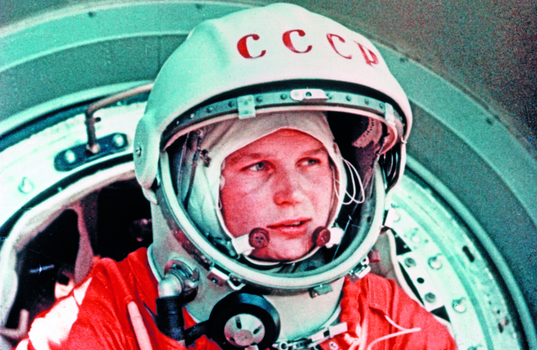 Fotografia. Retrato de uma mulher de olhos claros, vestindo capacete e traje laranja de astronauta em uma nave espacial.