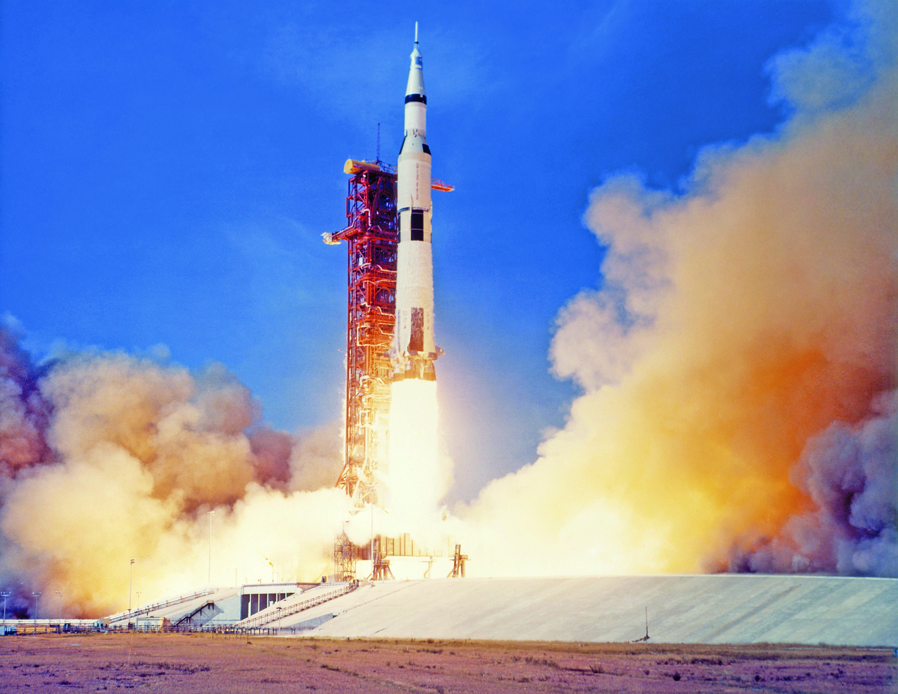 Fotografia. Foguete espacial em uma estação  no momento de seu lançamento, expelindo jatos de gás aquecido e fogo pela sua base, ladeada pela fumaça resultante desse processo.