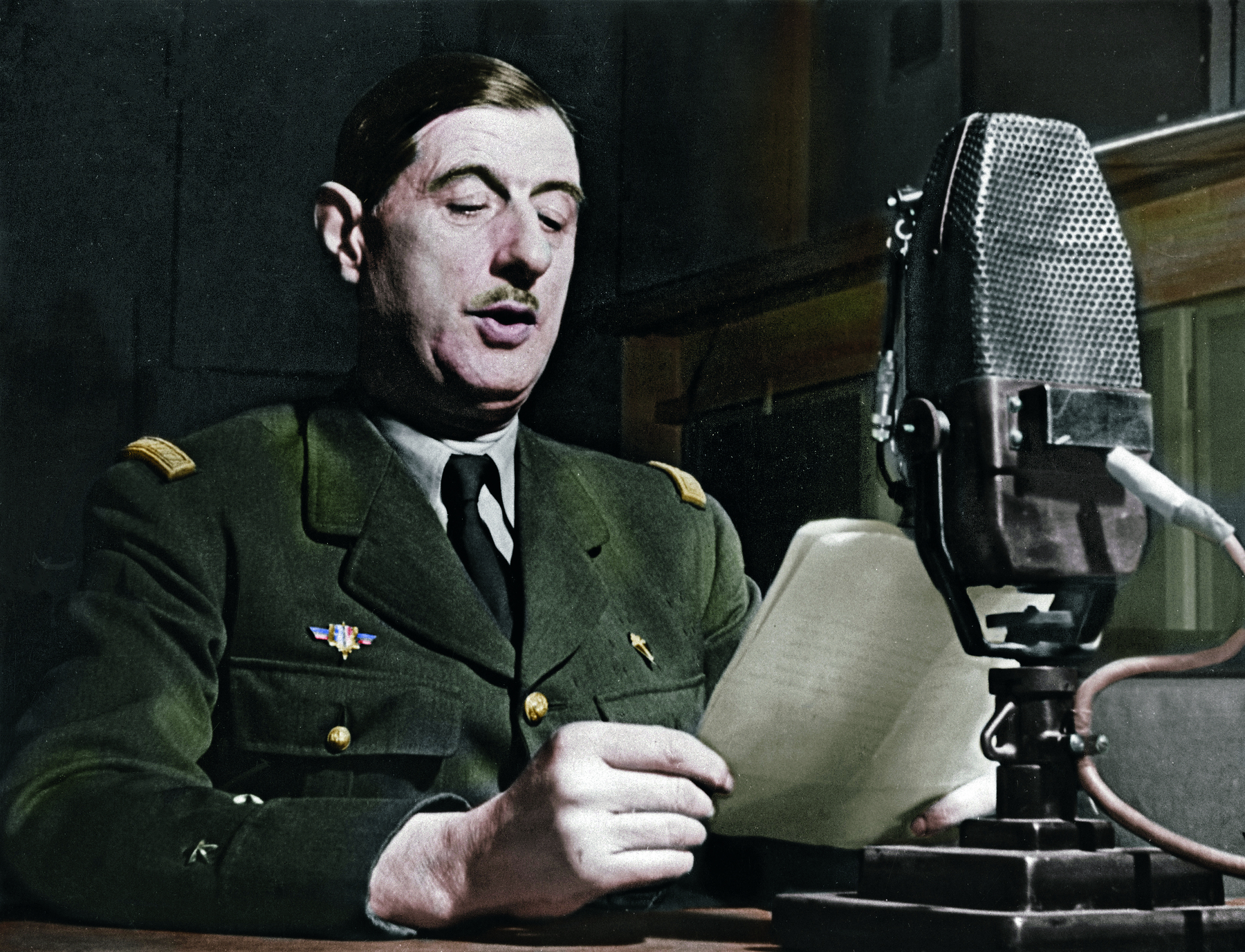 Fotografia. Um homem de cabelos castanhos, curtos e lisos, vestindo uma farda verde escuro, à frente de uma mesa contendo um microfone. Segura folhas de papel em suas mãos, voltadas para seu rosto.
