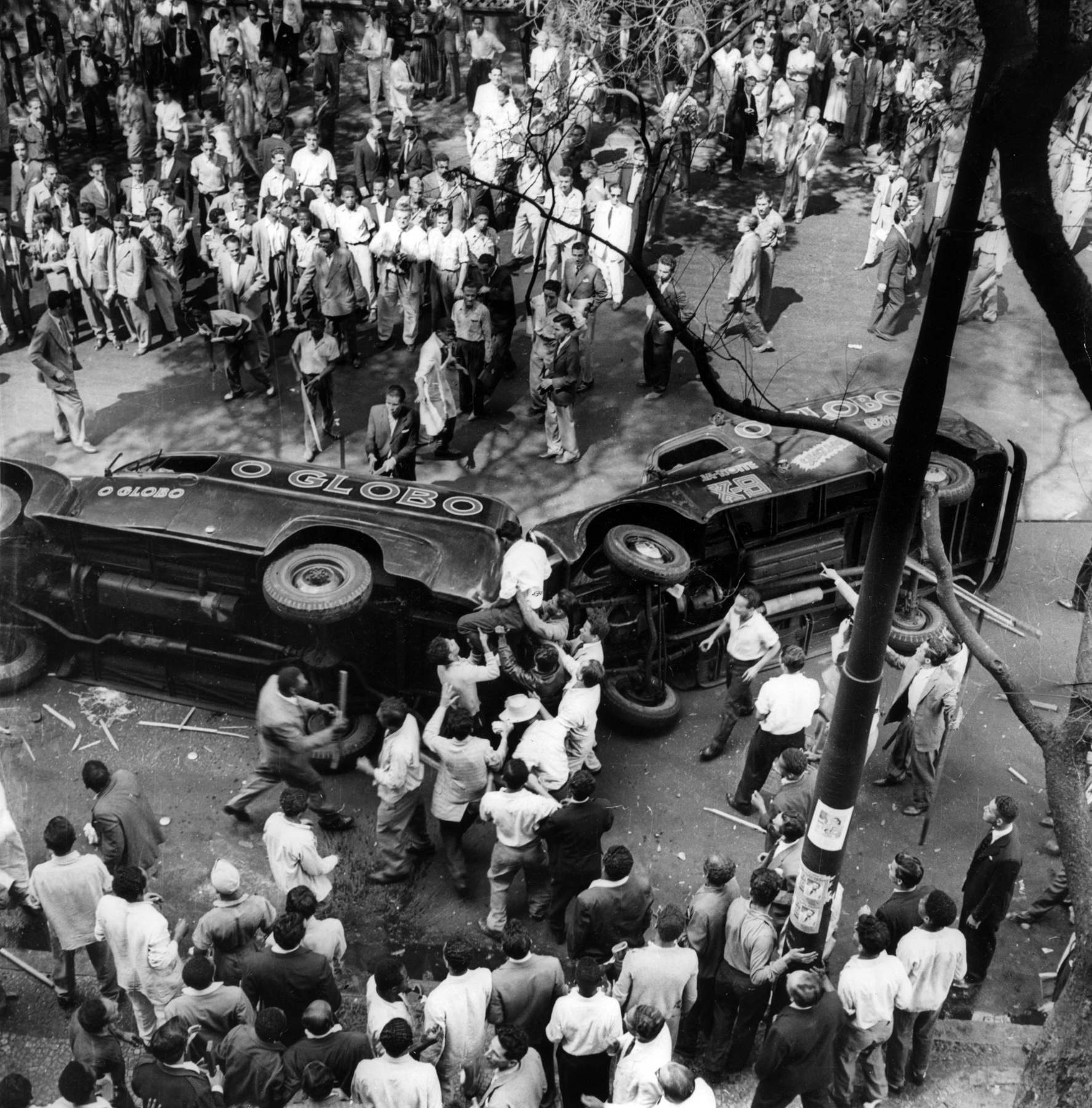 Fotografia em preto e branco. Dois veículos tombados, com as palavras 'O Globo' na lataria, e ao redor, dezenas de pessoas aglomeradas, algumas portando bastões.