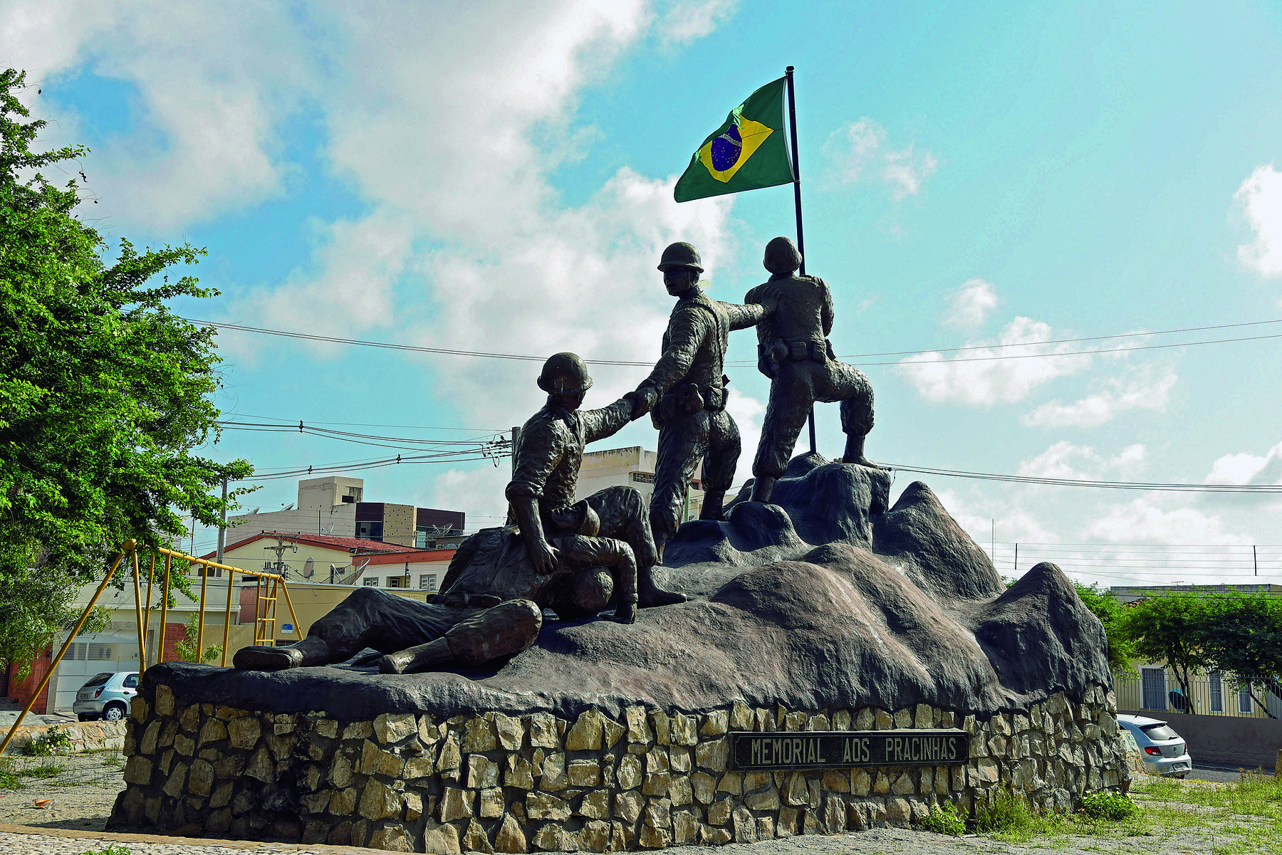 Fotografia. Destaque para um monumento cinza, representando um grupo de soldados sobre um rochedo irregular, direcionados a um mastro com a bandeira brasileira.