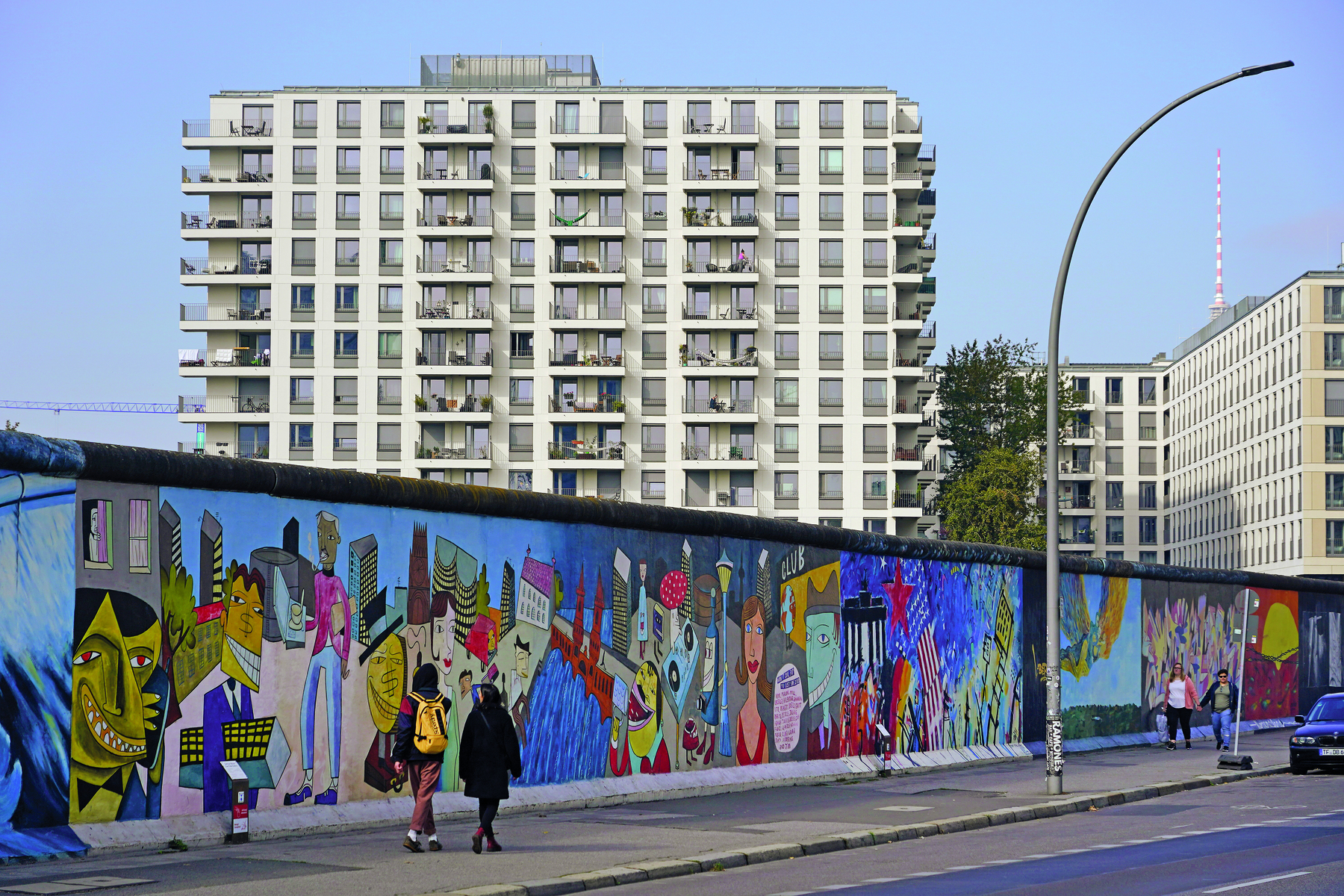 Fotografia. Em uma via, um muro extenso, repleto de grafites coloridos. Em segundo plano, prédios com vários andares, com sacadas e janelas distribuídas de forma homogênea pela fachada de paredes brancas.
